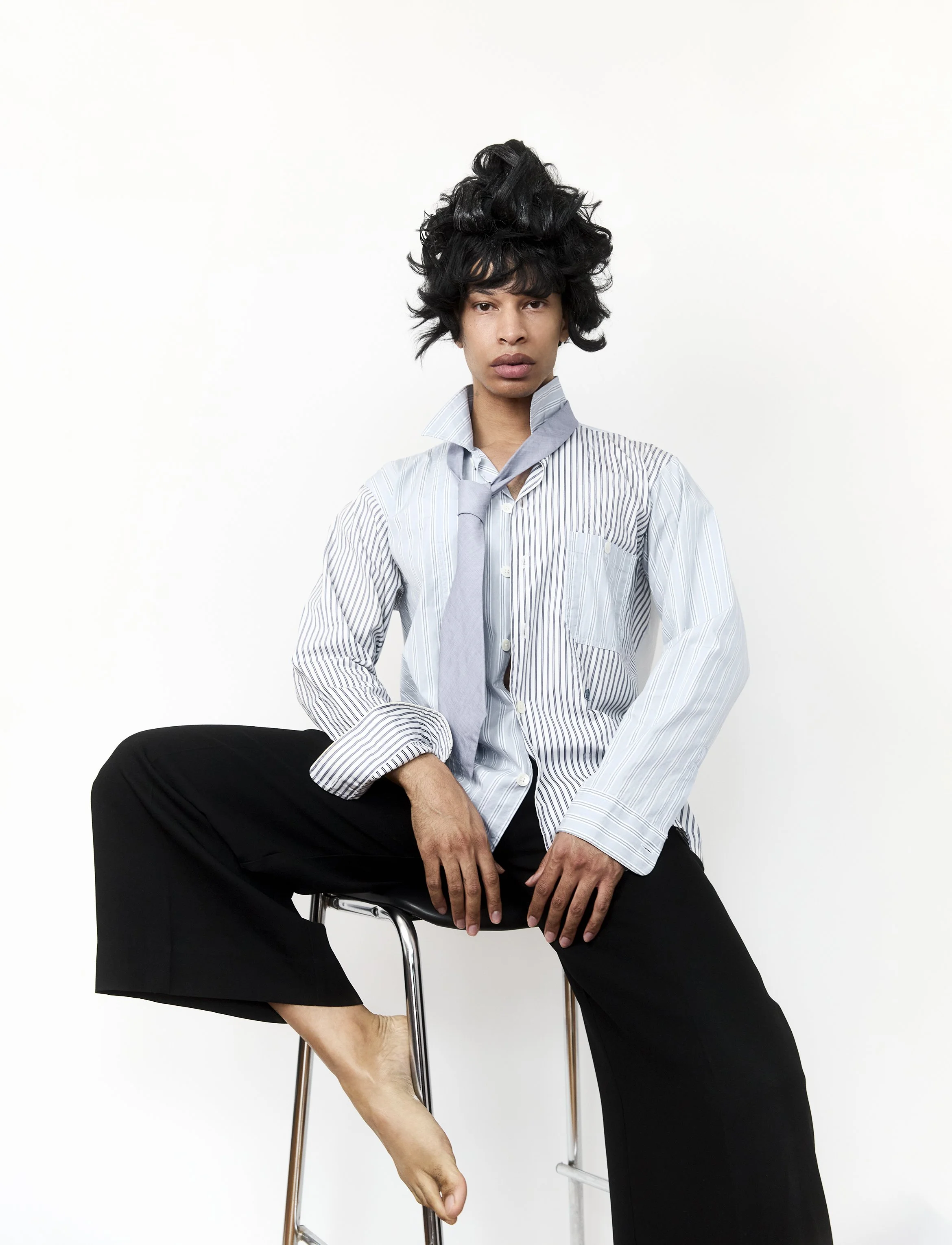 A person with dark, curly hair styled in an updo, wearing a light blue and white striped shirt with the top button undone, black pants, sitting on a metal stool against a plain white background.