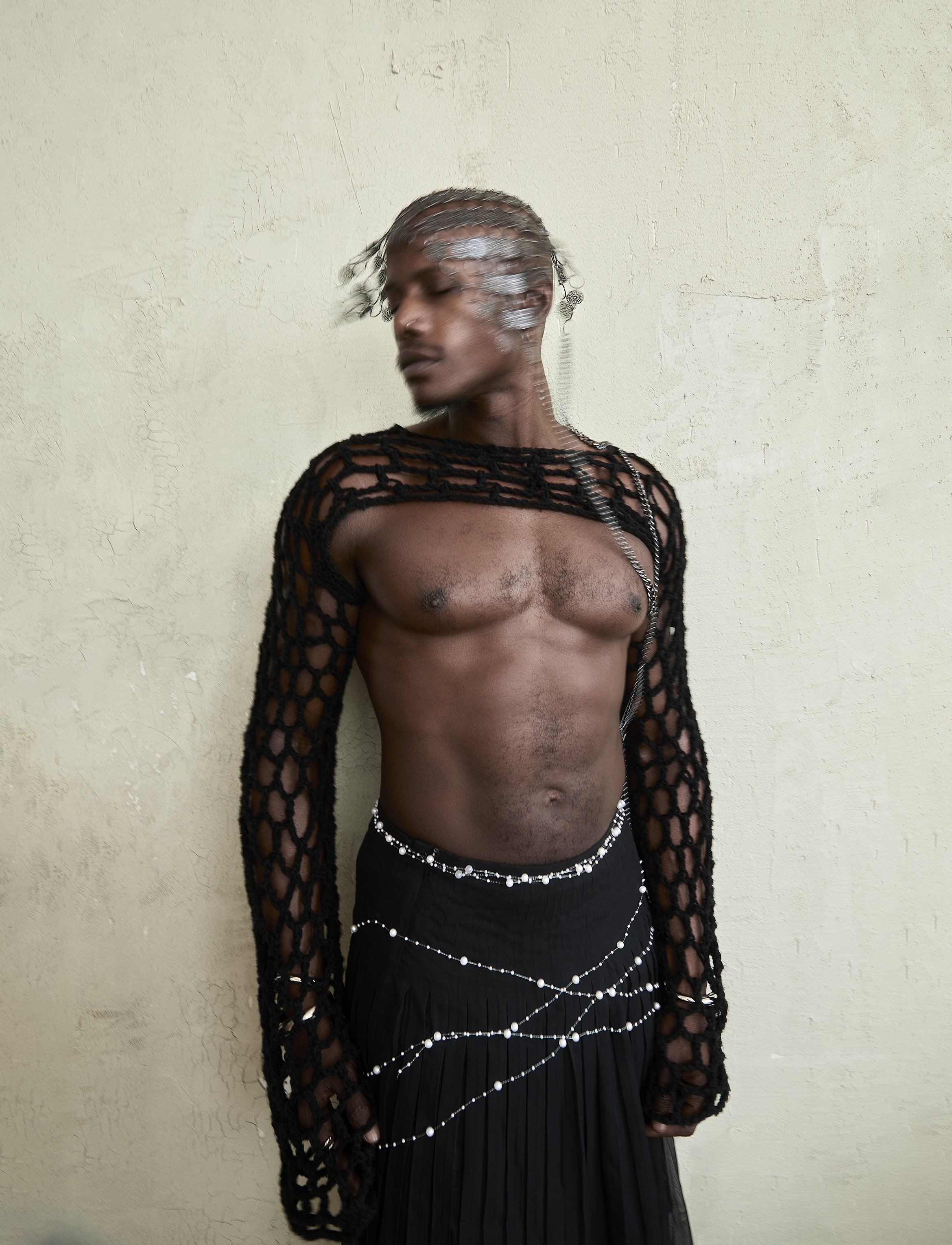 A shirtless person with short, curly silver hair standing against a textured white wall, wearing a black open-knit long-sleeve top and layered pearl necklaces.
