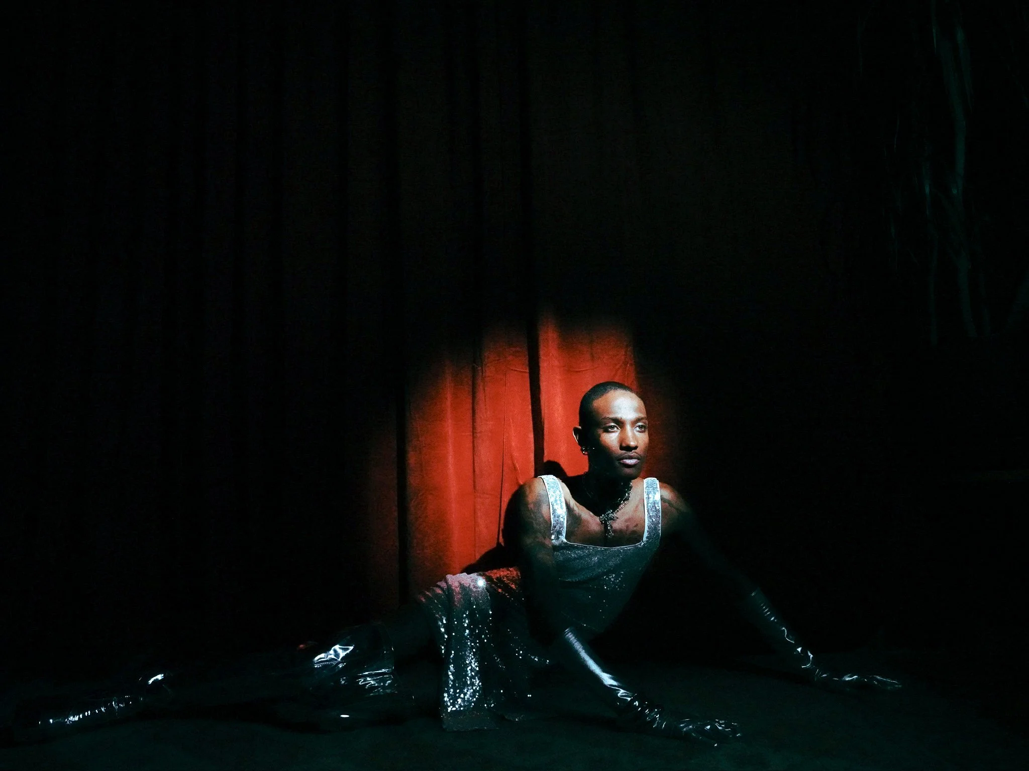 A performer on stage, dressed in a shiny, black outfit, with visible arms and head, stands in a spotlight against a dark background with a red curtain, during a theatrical or dance performance.