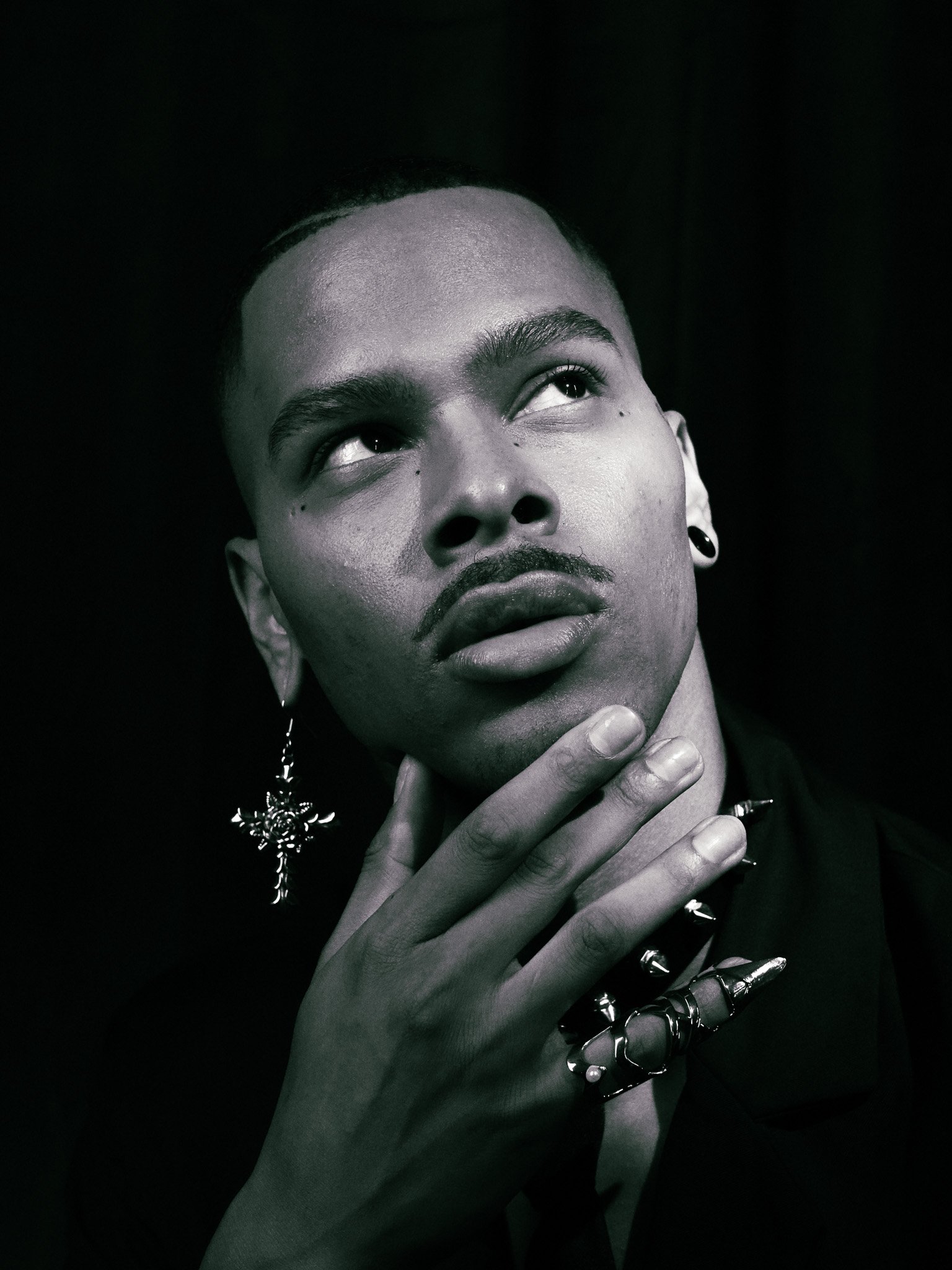 A person with short hair and earrings looking upwards, touching their face with one hand, in black and white.
