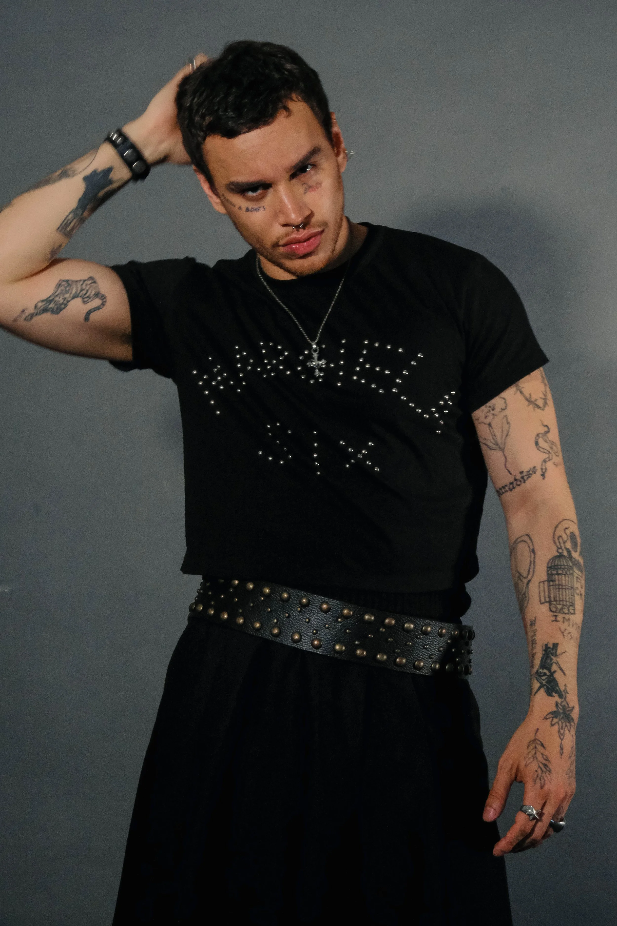 A young man with dark hair and tattoos poses against a plain wall, wearing a black T-shirt with rhinestone embellishments, a studded belt, and various jewelry, including rings, necklaces, and earrings.