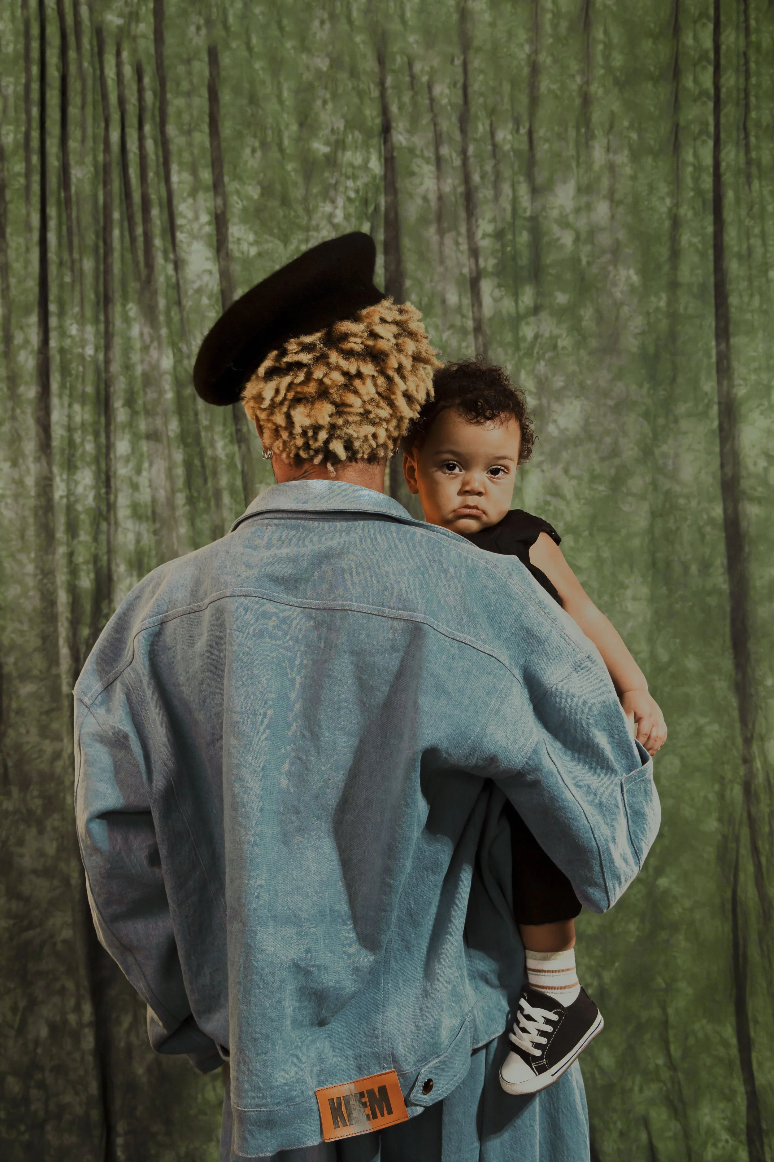A person with curly blonde hair wearing a denim jacket and a black hat is holding a young child with short curly hair, dressed in black with striped socks and sneakers, in front of a green, forest-like background.