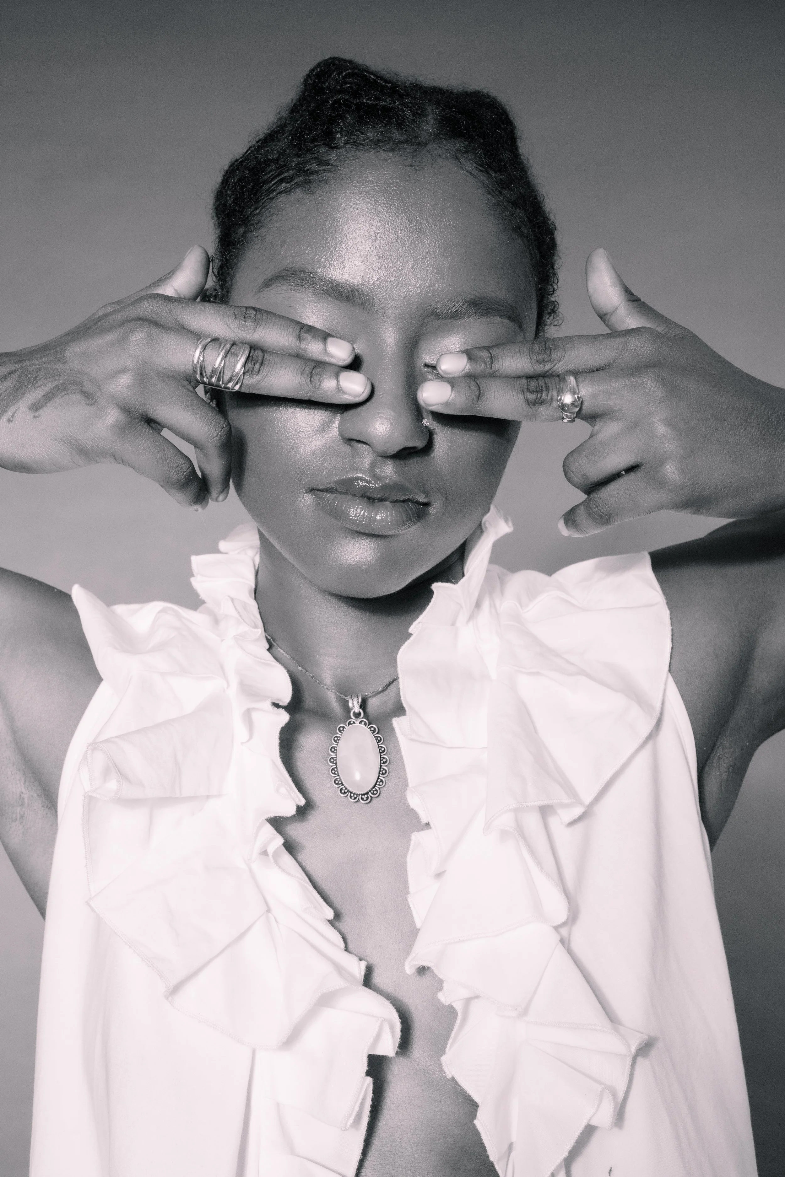 A woman with dark skin covering her eyes with her hands, wearing rings, a necklace, and a white ruffled top, in black and white.