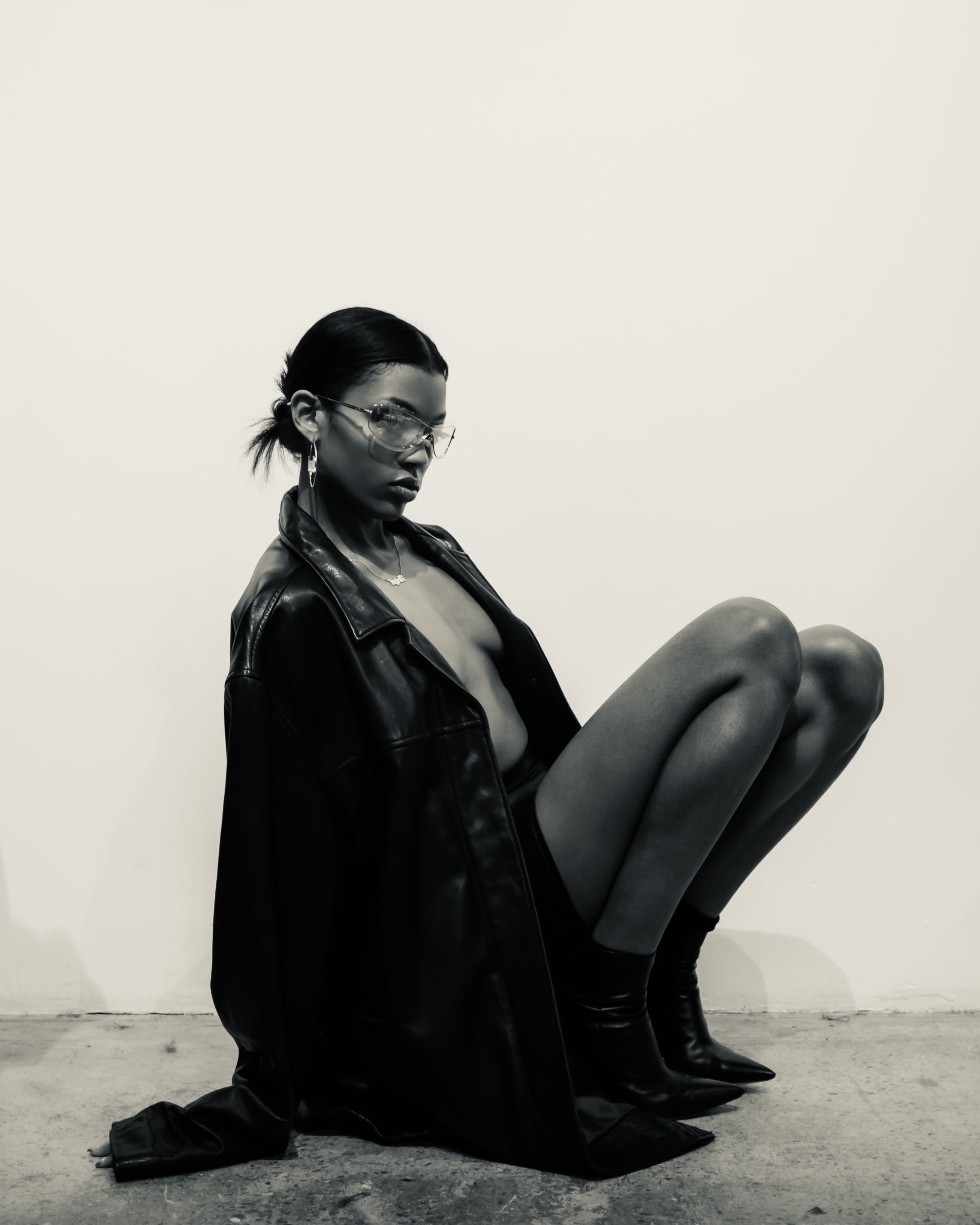 A woman with dark hair tied back, wearing sunglasses, earrings, a necklace, a leather jacket, and sitting on the floor of a minimalistic space, with her legs bent.