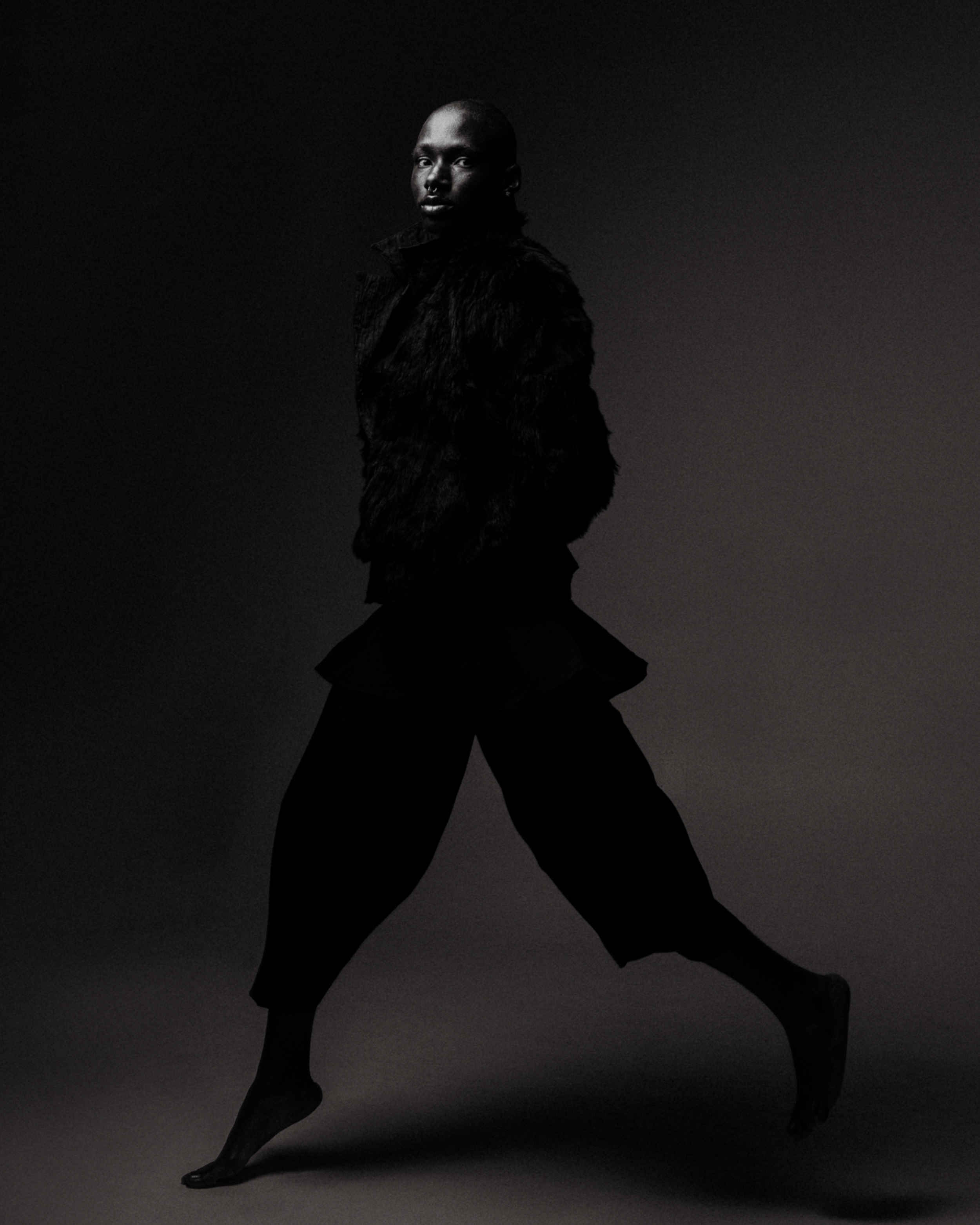 A person with a shaved head wearing a furry black jacket and black pants, posing against a dark background.
