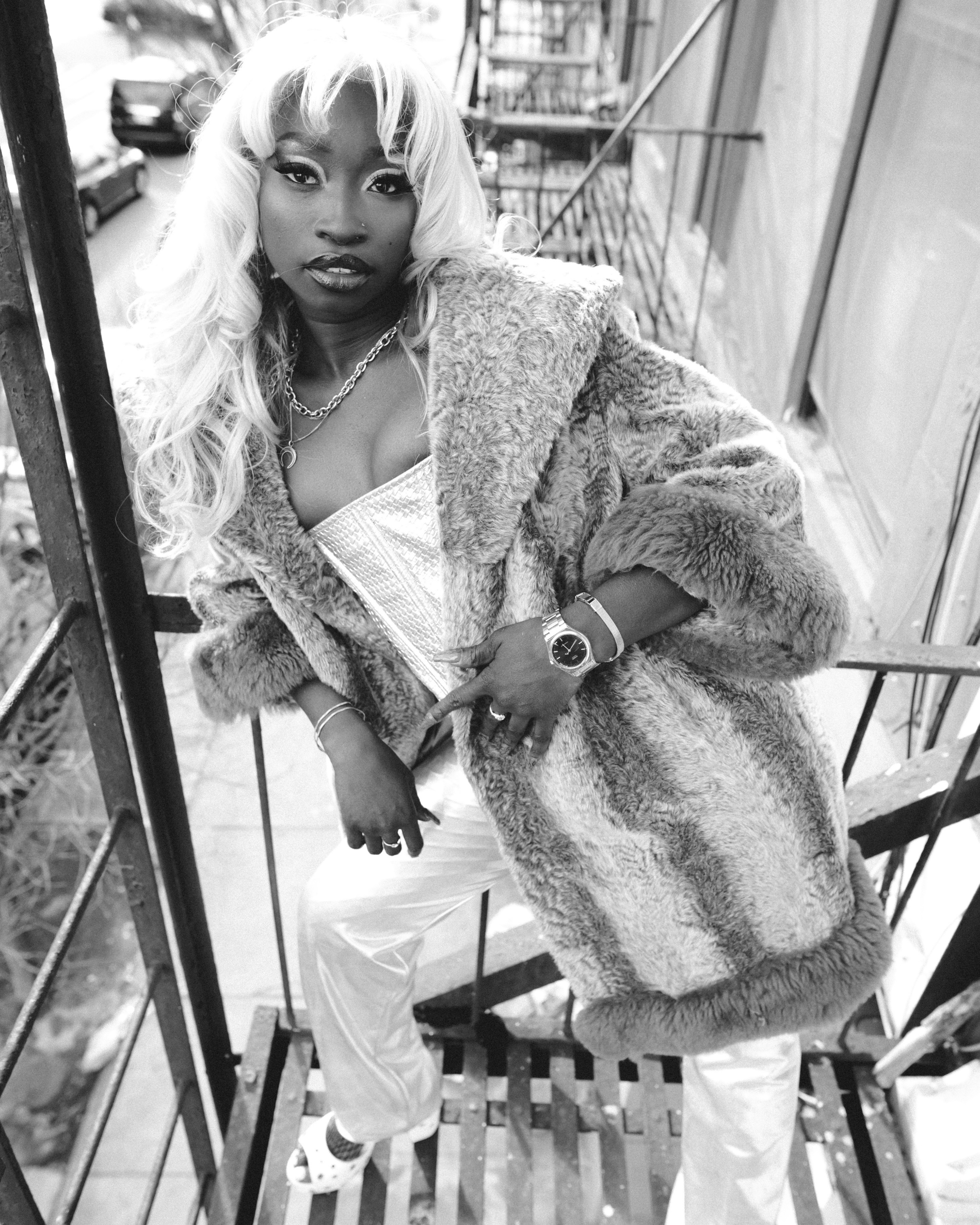 Black-and-white photo of a woman with light-colored, wavy hair wearing a fur coat, standing on a porch or balcony with a metal railing. She is leaning slightly against the railing with one arm crossing her body and her hand resting on her waist. She 