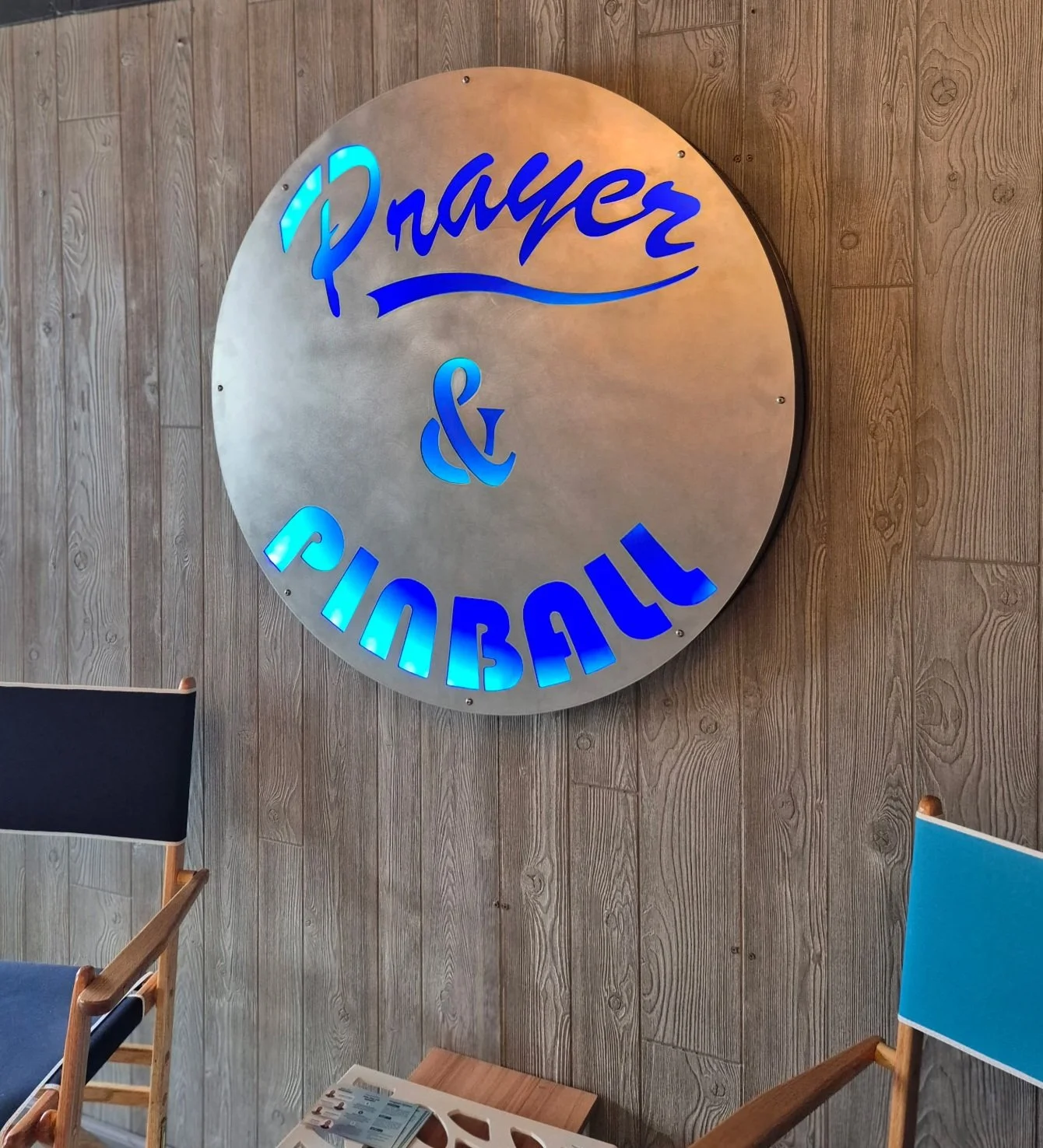 Custom fabricated metal sign reading "Prayer & Pinball" which has LED lighting. The sign is round and silver colored.
