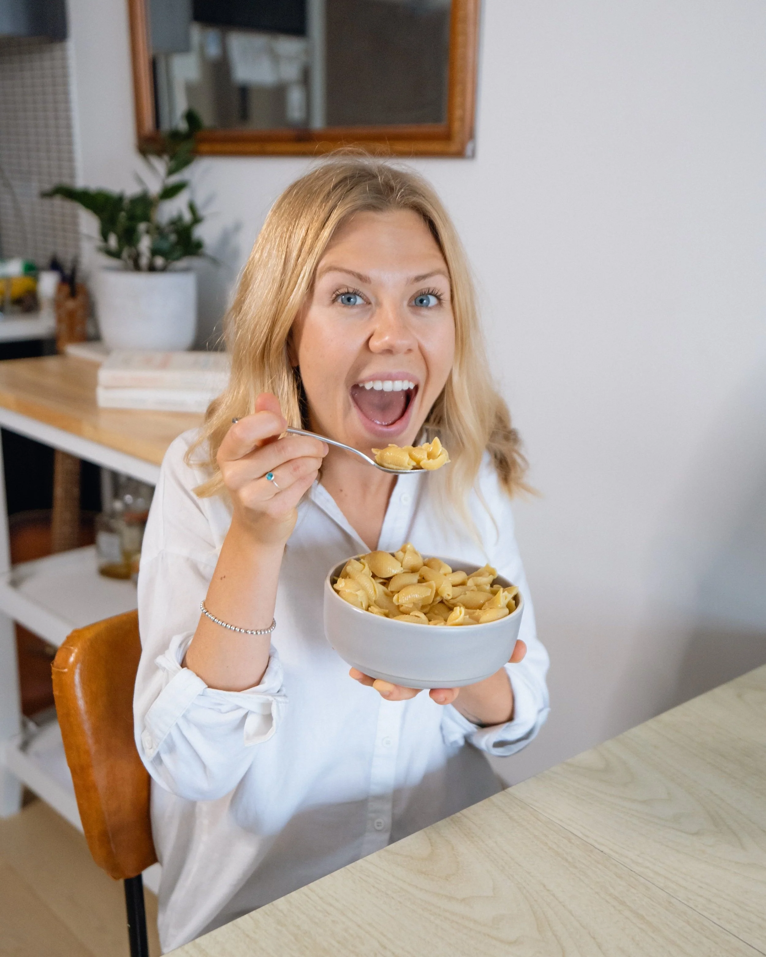 A woman with blonde hair and blue eyes sitting at a wooden table, holding a bowl of pasta in one hand and a spoonful of pasta in the other, smiling excitedly.