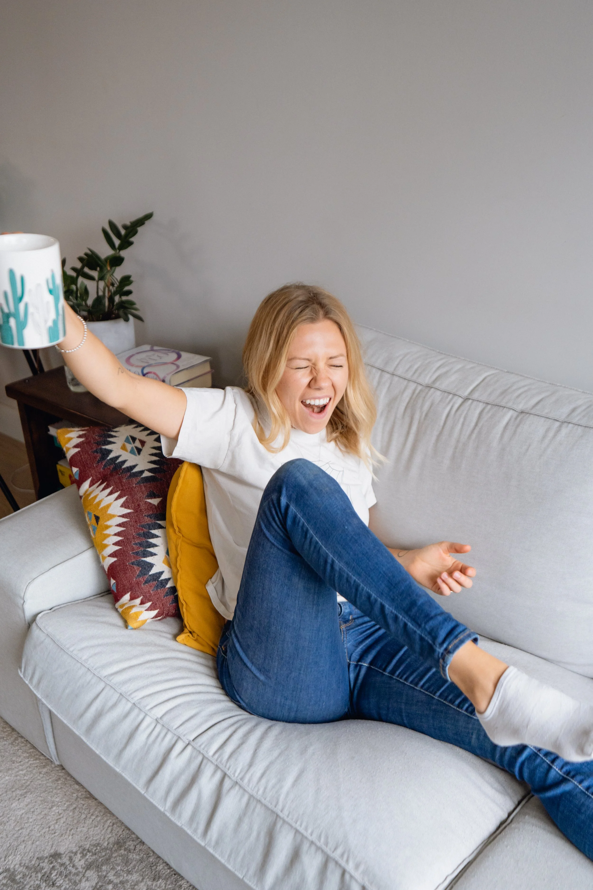 A woman sitting on a white couch, laughing with her eyes closed, holding a mug with a cactus design in her right hand, and wearing a white t-shirt and blue jeans.