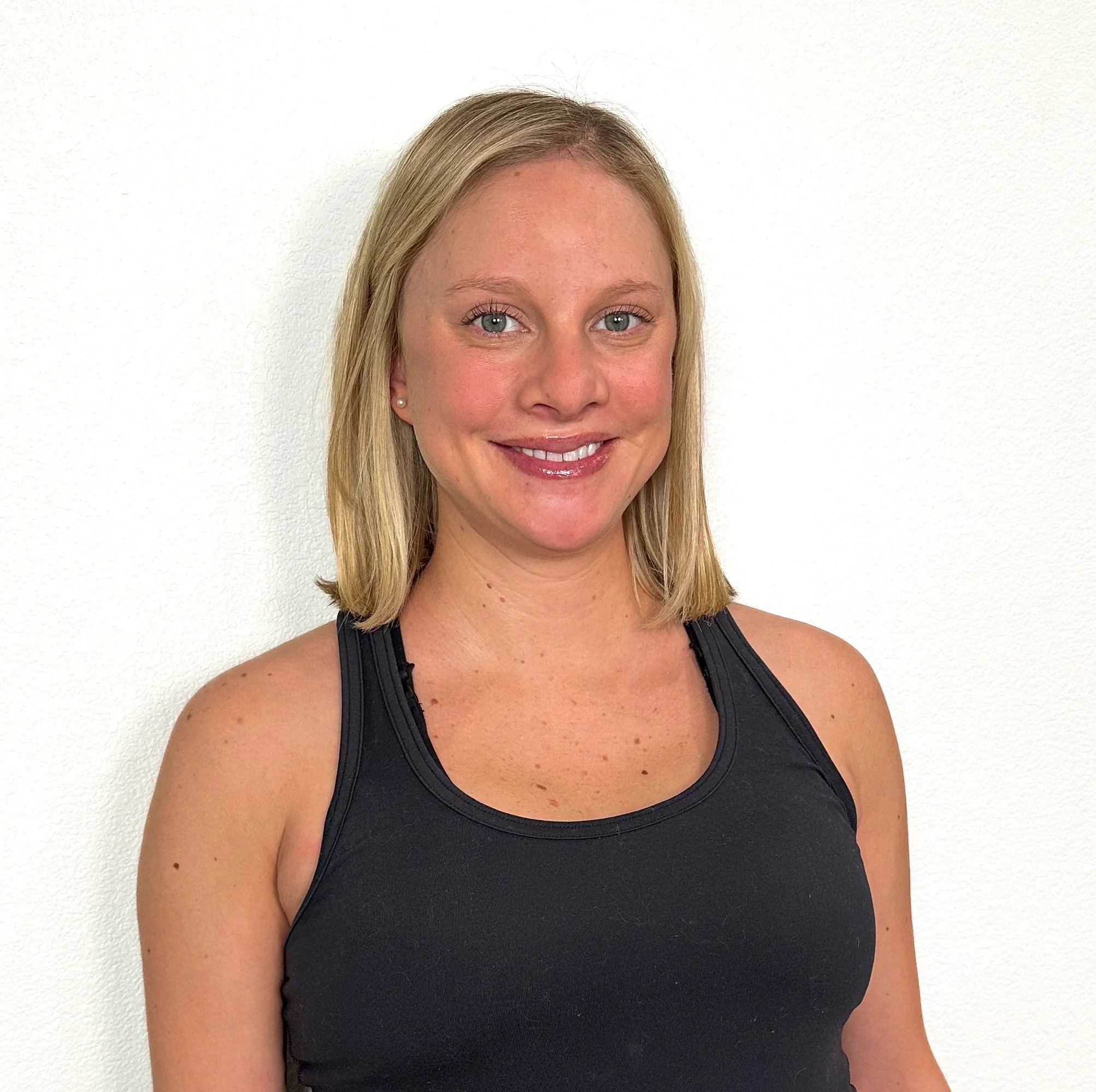 A woman with blonde hair, wearing a black athletic top, smiling, standing against a plain white wall.