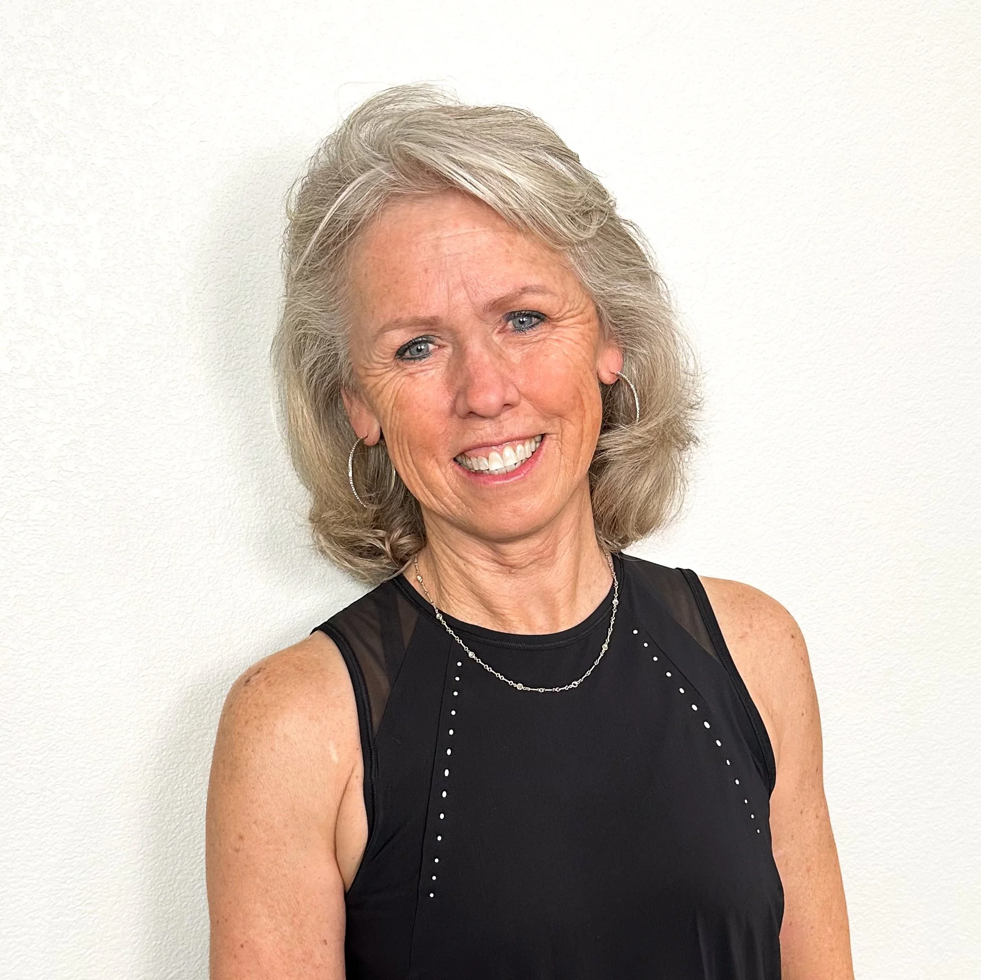 Smiling middle-aged woman with shoulder-length blonde hair, wearing hoop earrings, a necklace, and a black sleeveless top, standing against a plain white wall.
