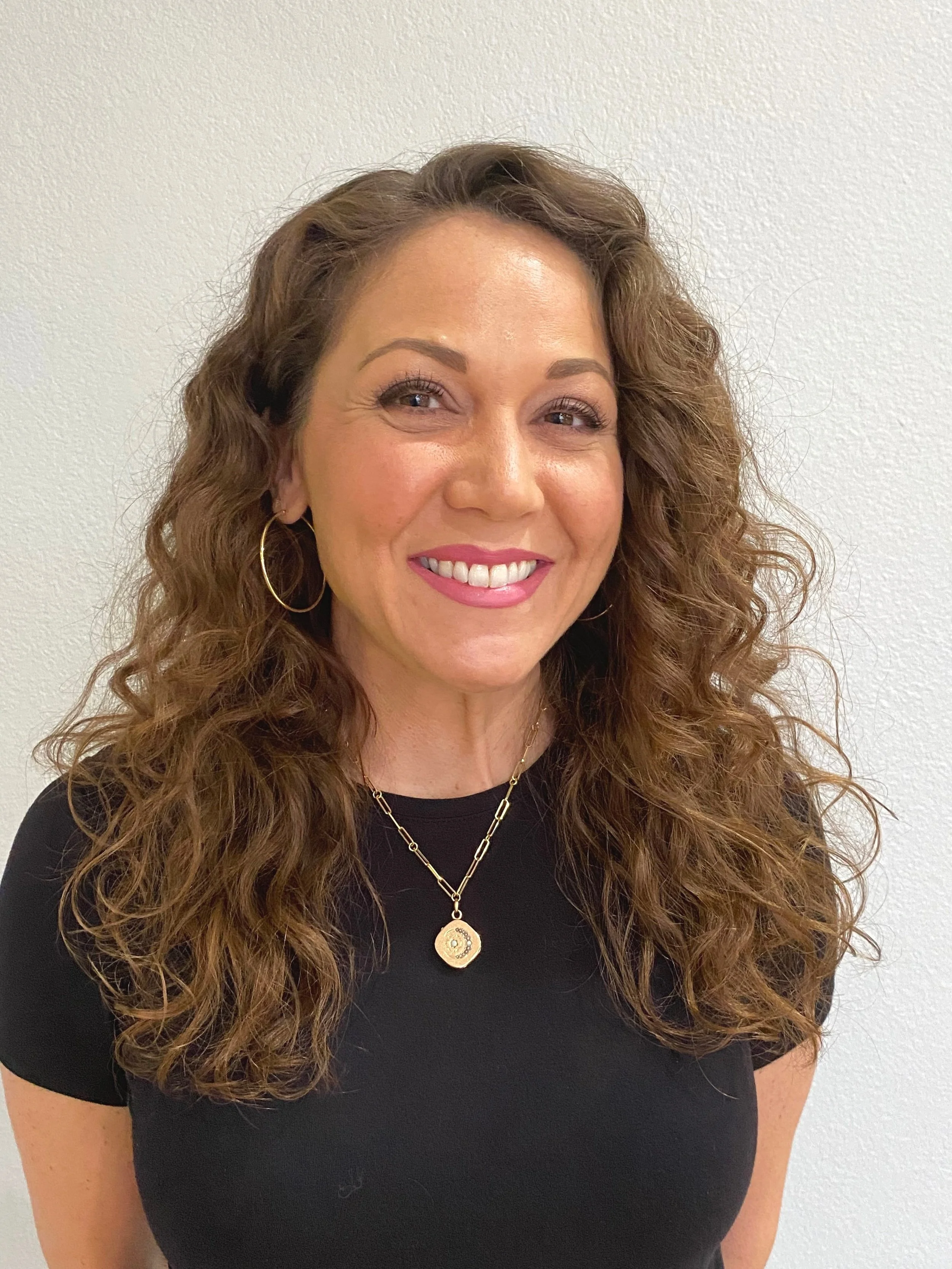 A smiling woman with long, curly brown hair wearing a black top, gold hoop earrings, and layered gold necklaces, standing against a plain light-colored wall.