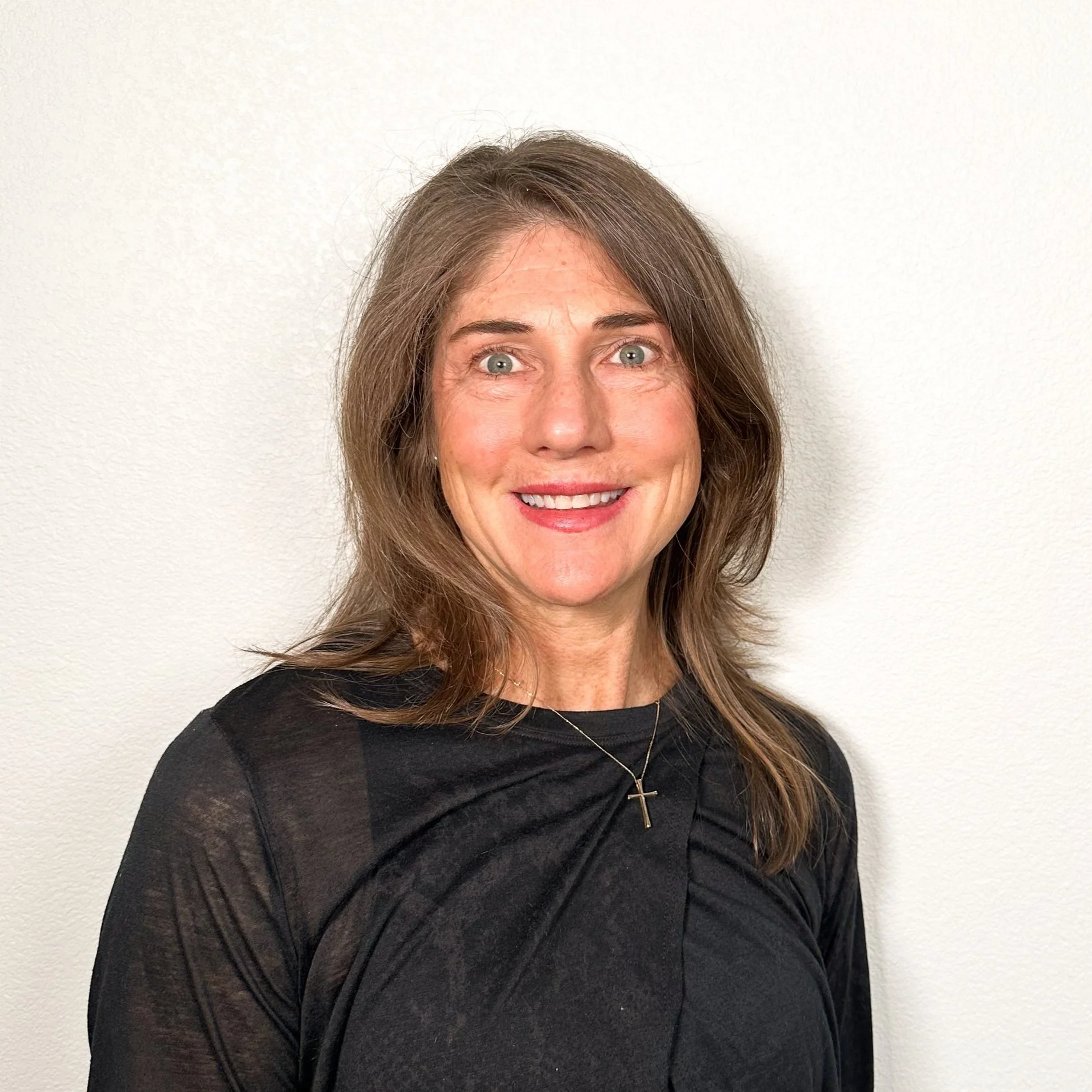 A woman with shoulder-length brown hair, blue eyes, and a cross necklace, wearing a black top, smiling in front of a white wall.