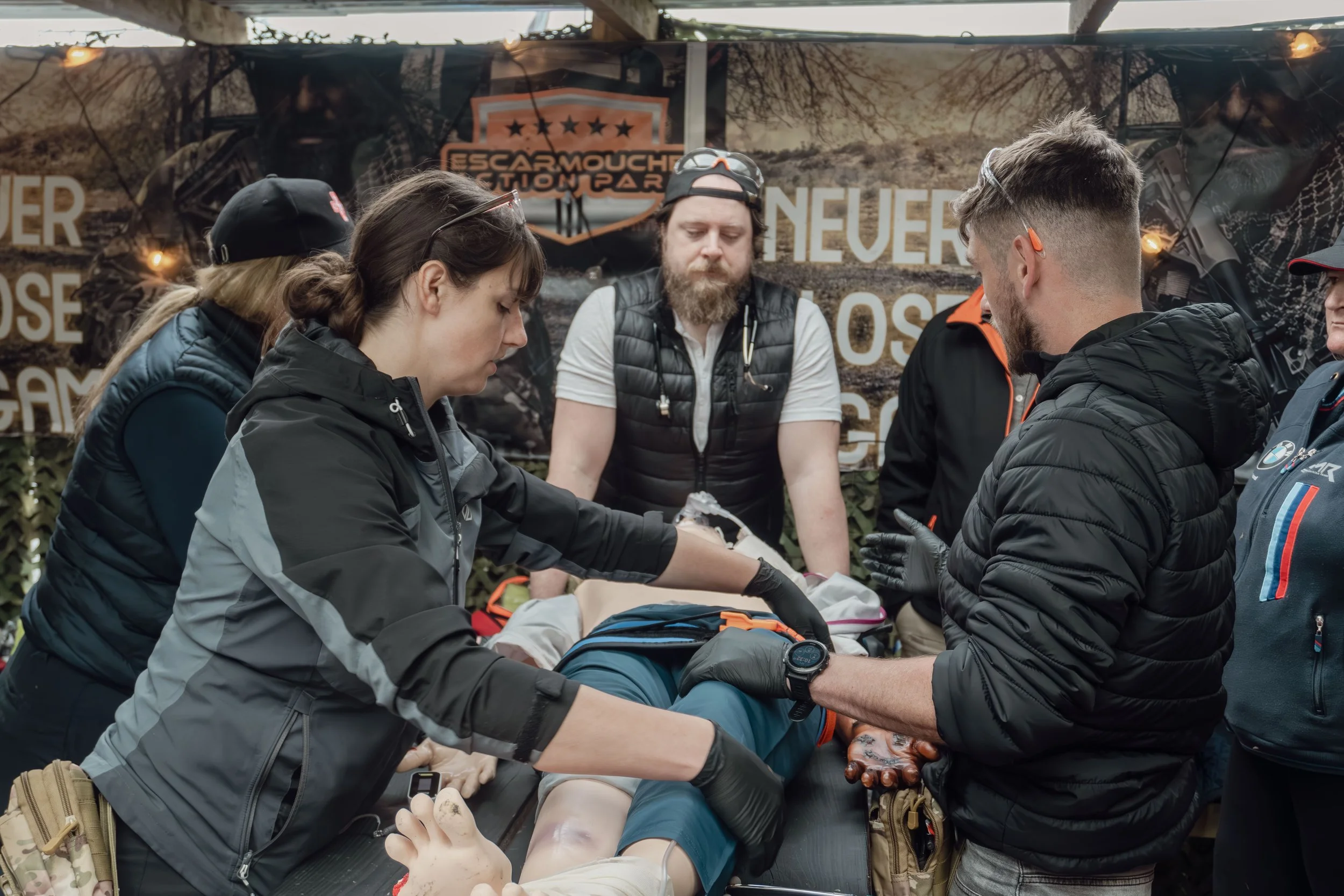 Group of people practicing emergency medical procedures on a person lying on a stretcher at an outdoor event with a banner in the background.