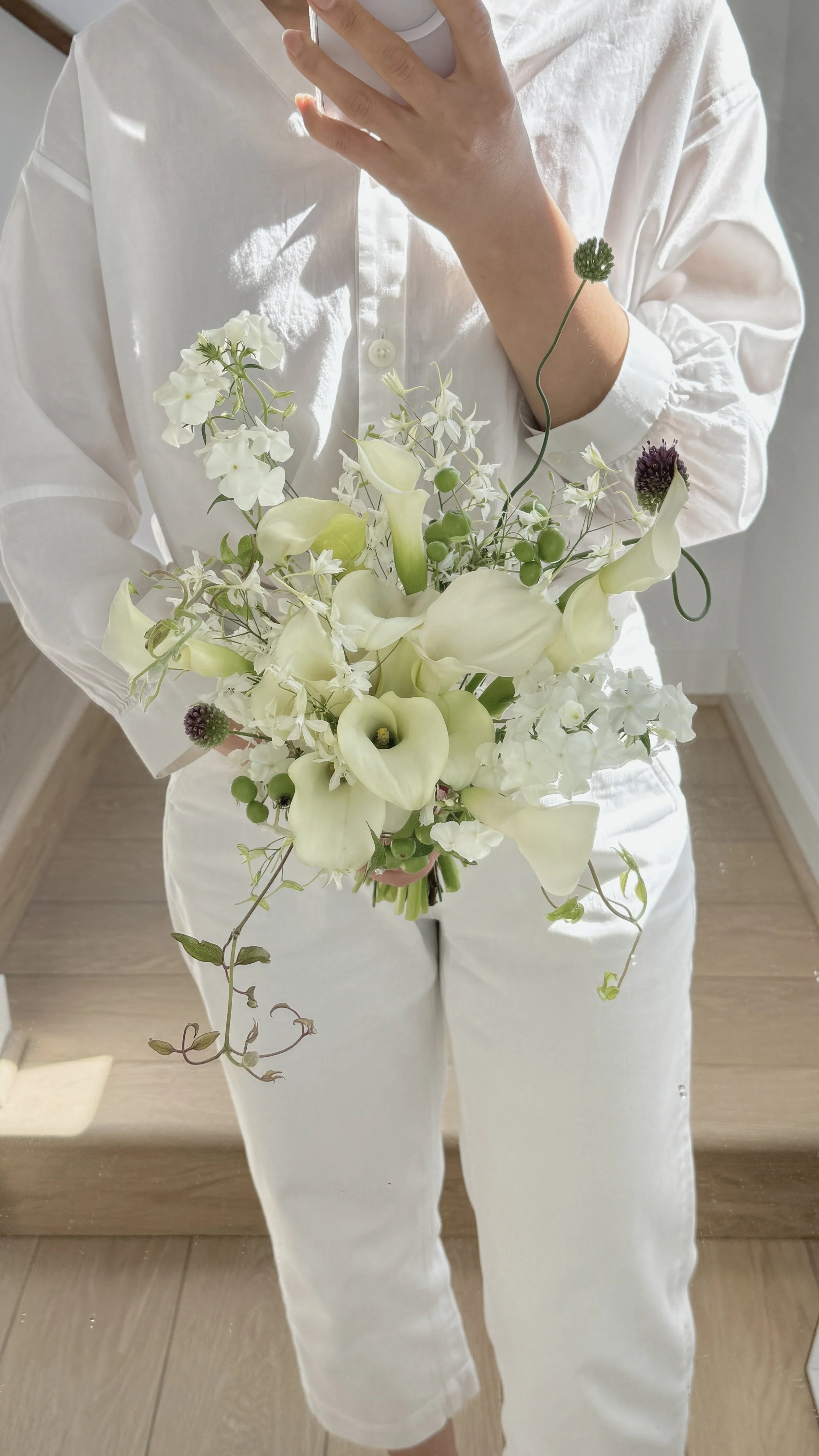 Person in white clothes holding a bouquet of white flowers, taking a mirror selfie.