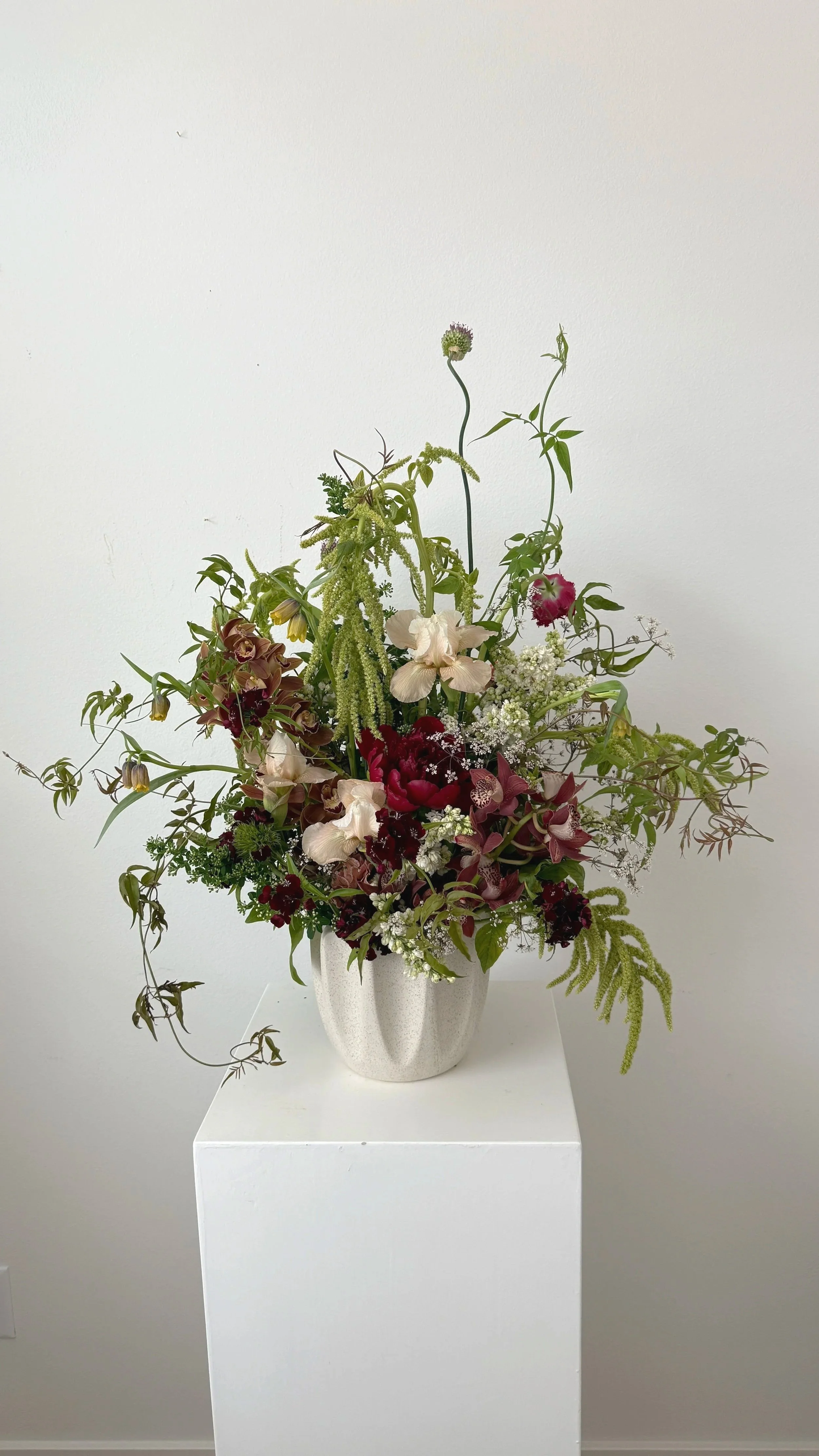 A floral arrangement in a white textured ceramic vase on a white pedestal against a plain white wall. The arrangement includes various green and burgundy flowers and foliage with some delicate white accents.