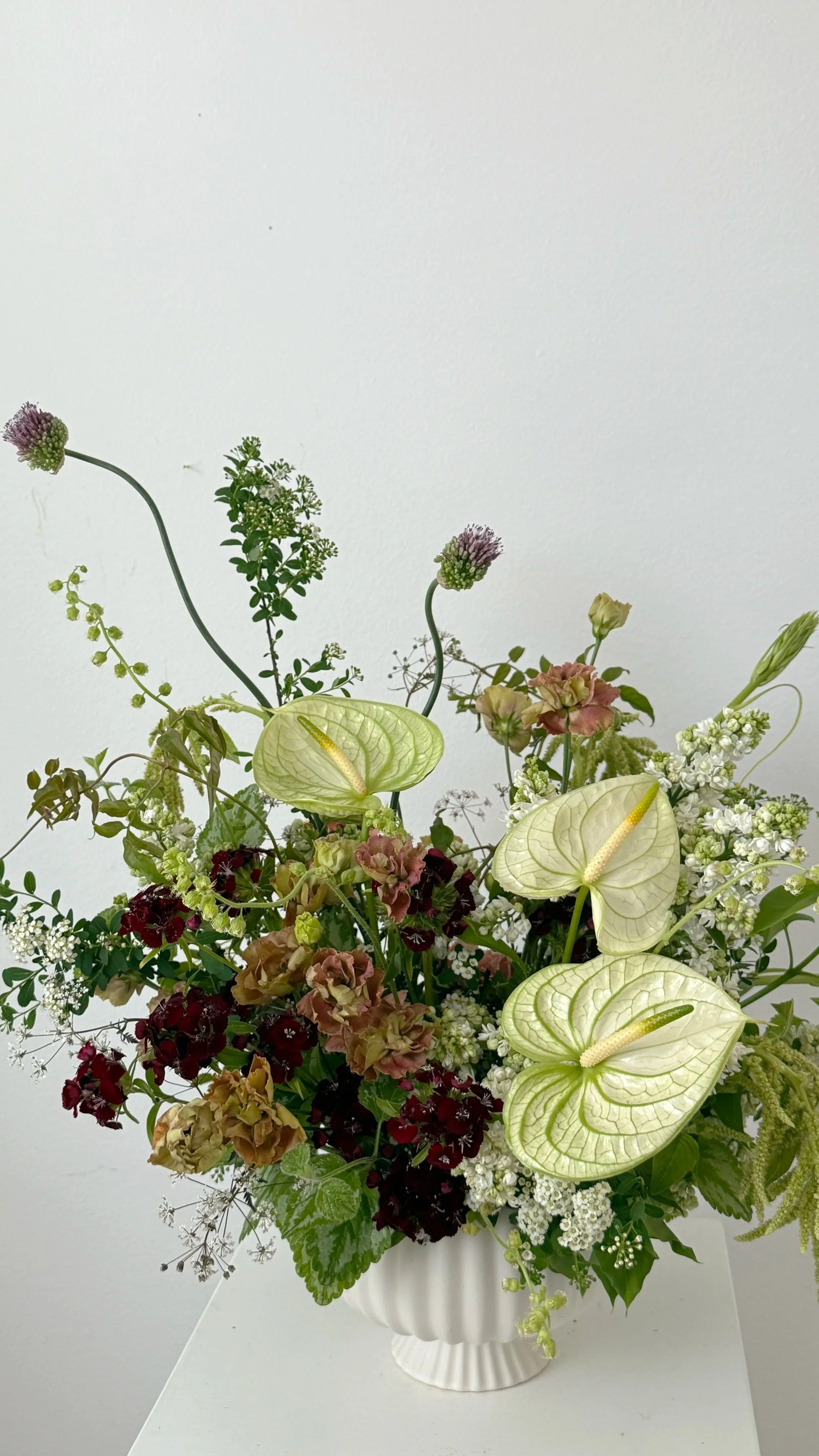 A floral arrangement in a white vase with an assortment of flowers, including anthuriums, white lilacs, and other white, pink, and dark red flowers, set against a plain white background.