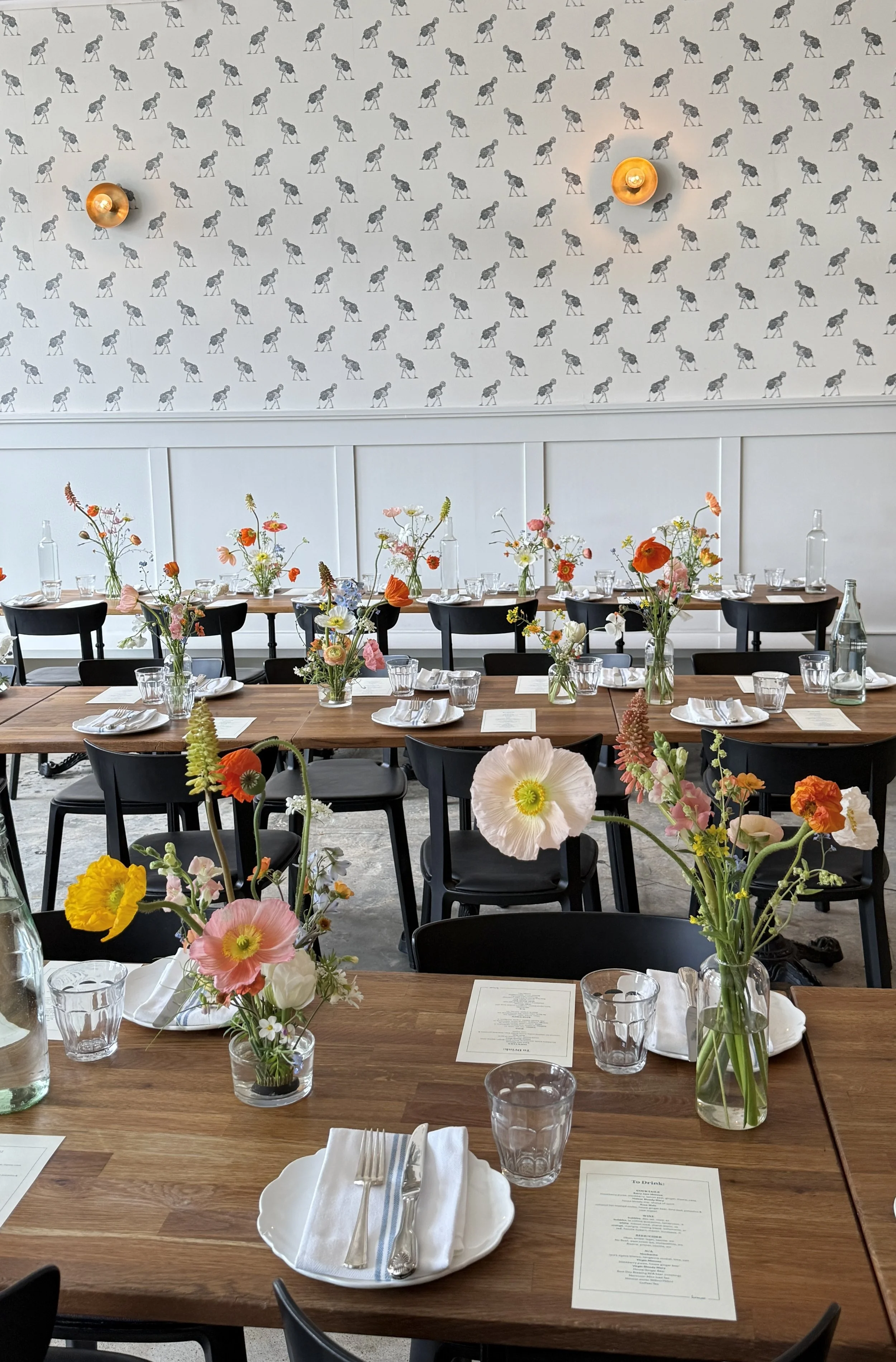 A dining setup with a long wooden table decorated with colorful flower vases, white plates, silverware, and glasses, in a room with a wall covered in black and white zigzag patterned wallpaper and a ceiling-mounted light fixture.