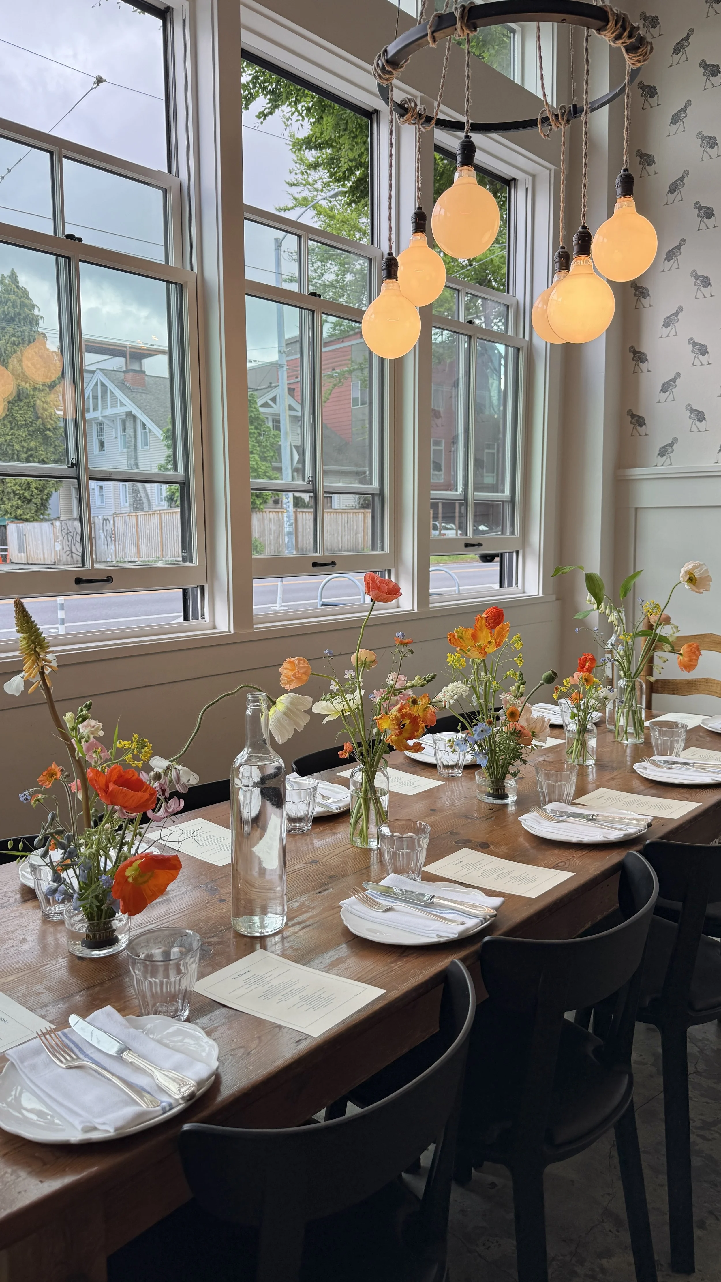 Interior of a dining room with a long wooden table set for a meal, decorated with colorful flower arrangements in vases, white plates, glassware, and menus. Large windows with multiple panes let in natural light and reveal a view outside of houses an