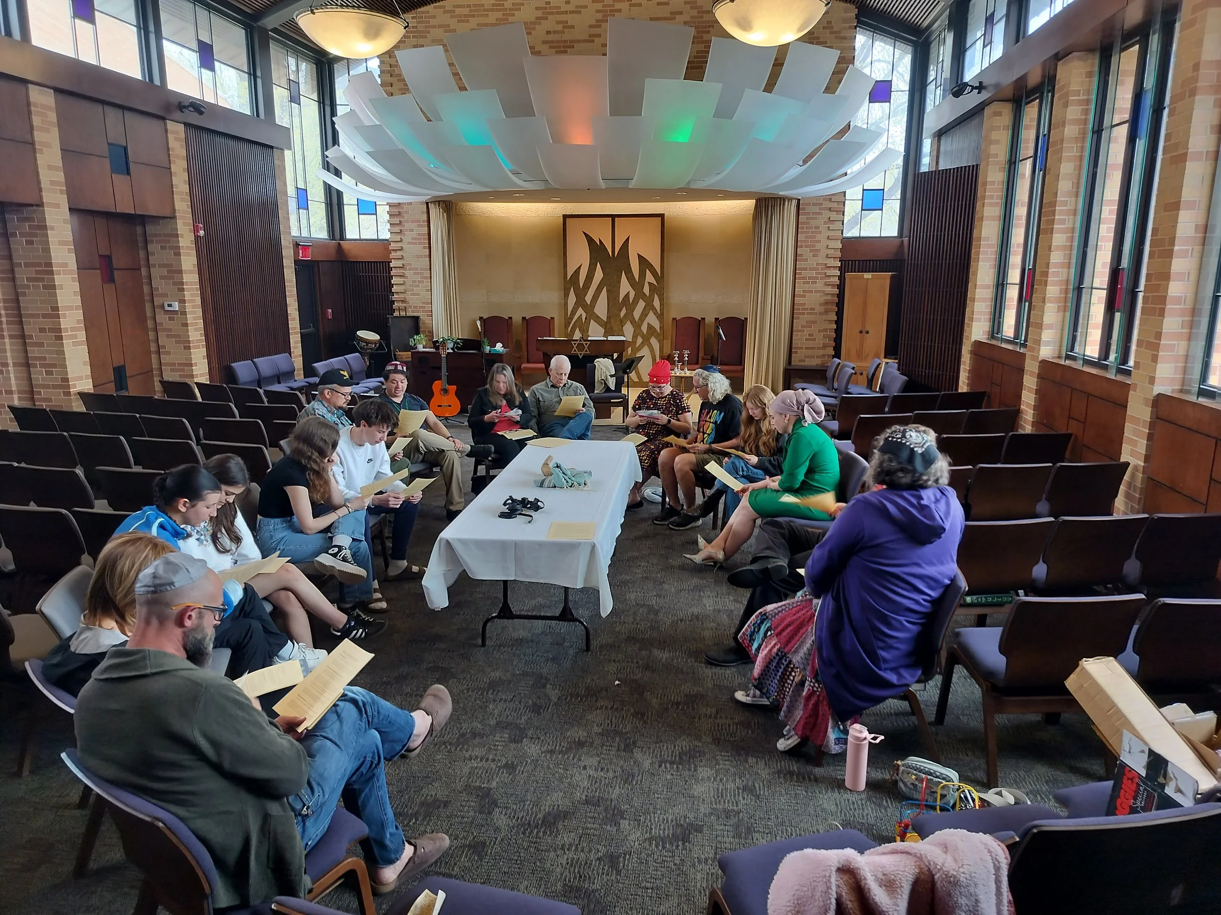 A group of people sitting in a circle inside a church or community center, reading papers or books, with musical instruments like a guitar and piano in the background, and a table in the center with a cloth and some objects.