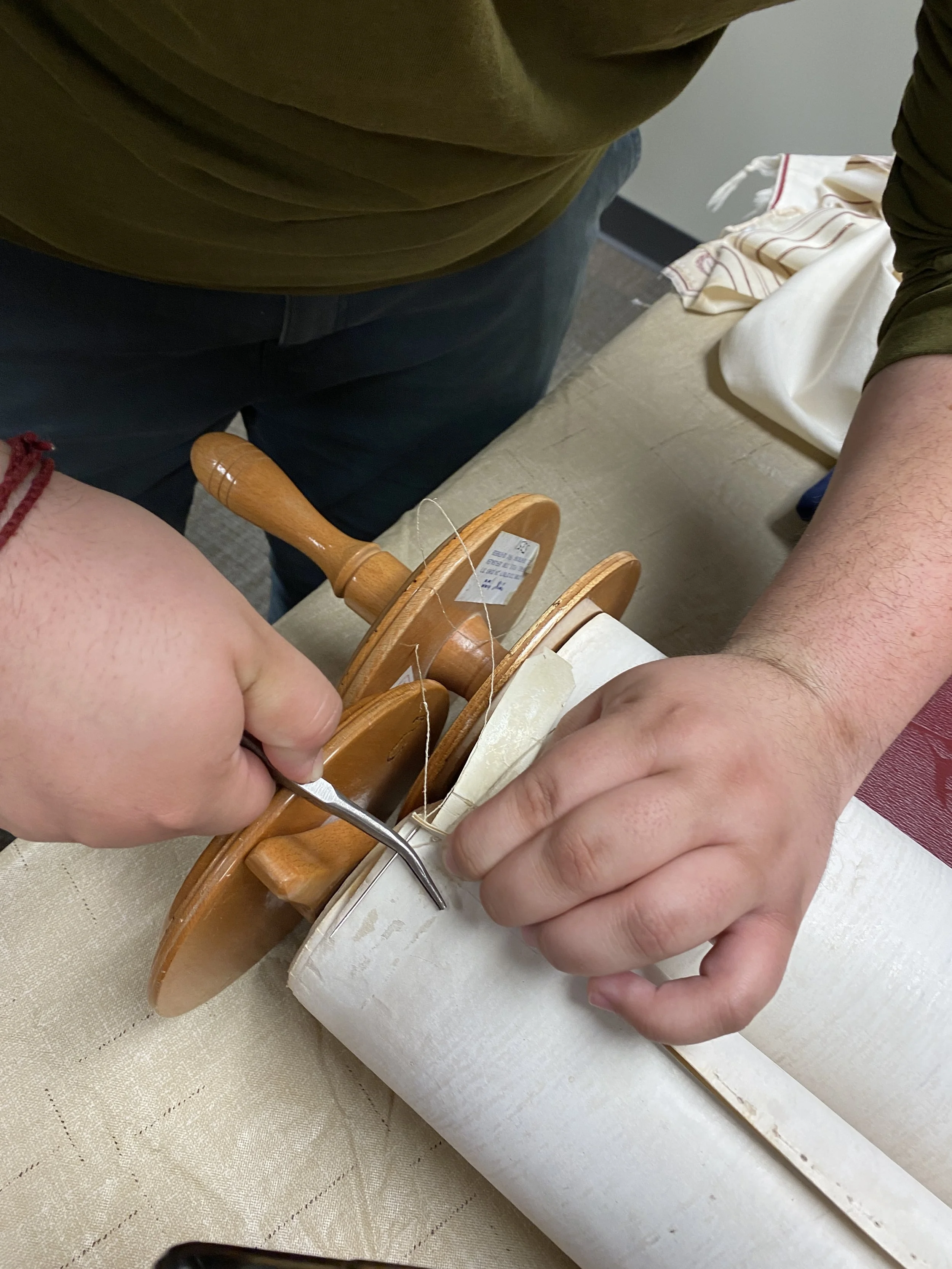 Two people are working together on a craft project involving a white ceramic object and a wooden wheel. One person uses a tool to attach something to the ceramic, while the other holds the object steady.