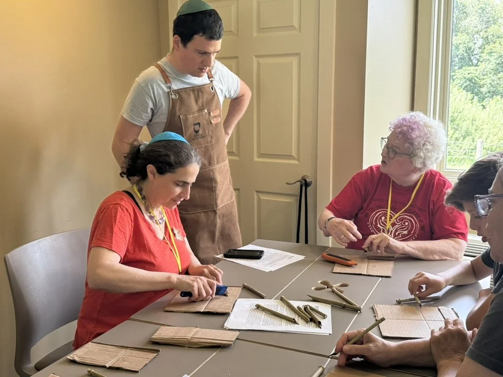 A group of older women and a young man are seated at a table engaging in arts and crafts. The women are wearing red shirts and safety glasses, and the young man, standing behind them, is wearing a brown apron. The table has wooden sticks, scissors, and papers on it. The background shows a window with a view of trees and a yellow wall.