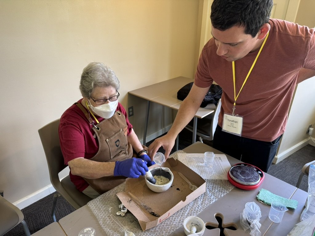 A woman wearing glasses, a face mask, an apron, and gloves is sitting at a table while a man with a name badge pours something into a bowl in front of her.