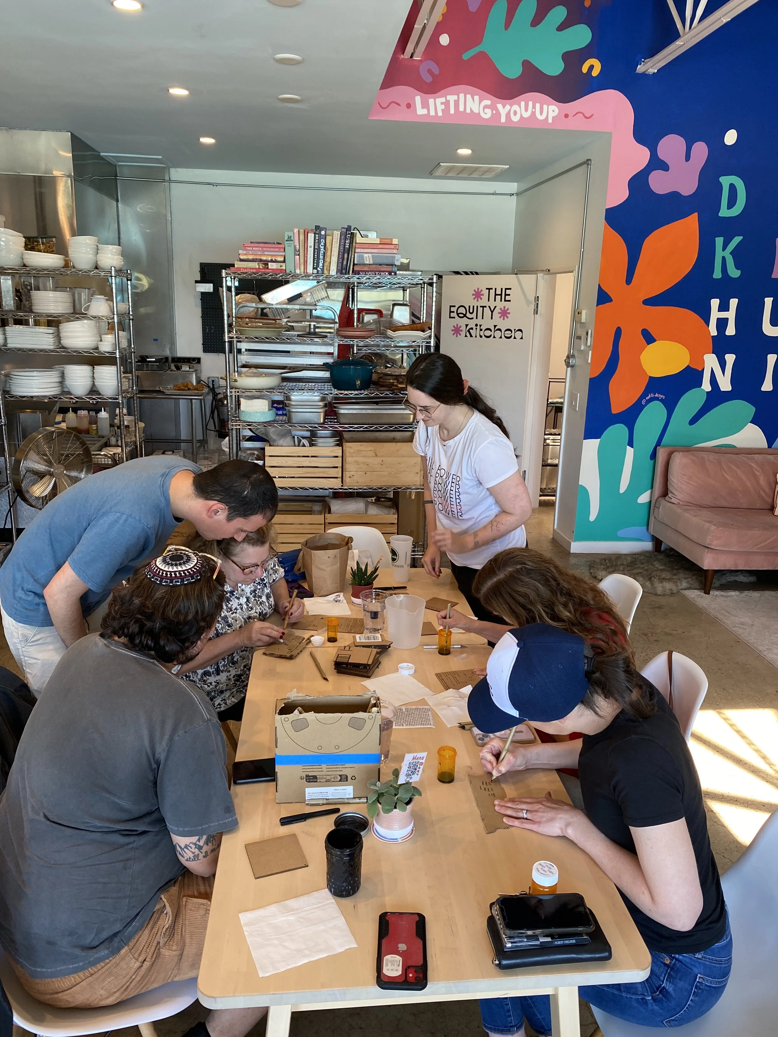Group of people sitting around a wooden table, engaging in a craft activity, in a well-lit room with colorful wall art and shelves filled with dishes and books.