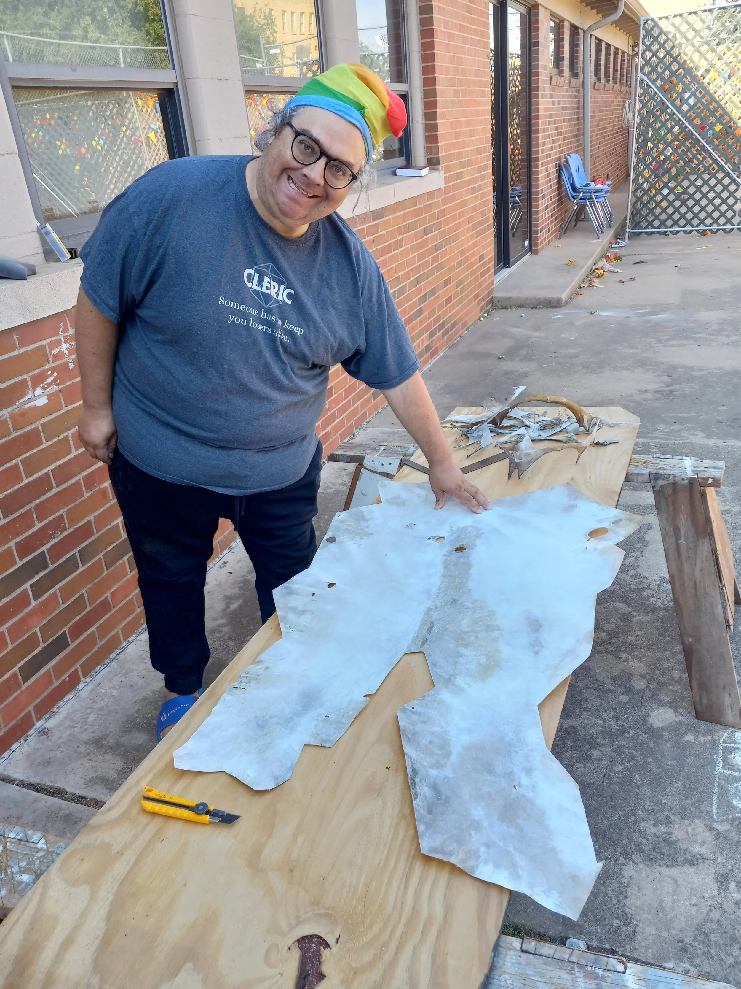 A person wearing glasses, a blue T-shirt with the words 'CLEPIC Someone has to keep you losers alive.', and a rainbow-colored hat, is standing outside on a concrete surface. They are pointing at a large piece of metal on a wooden worktable. There are various tools and smaller pieces of metal nearby, with a brick building and some chairs in the background.