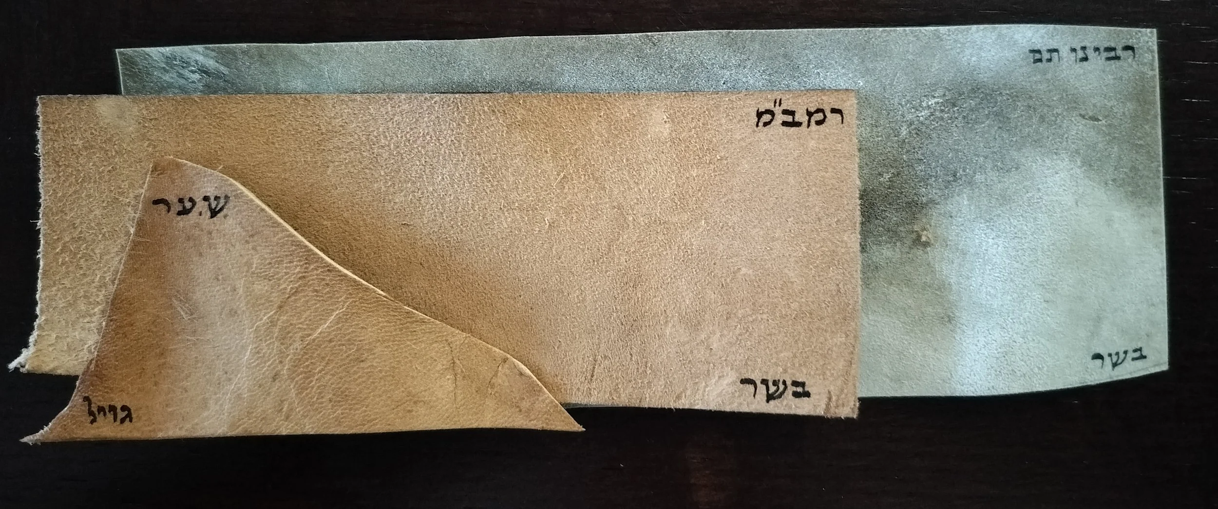Layered pieces of paper with edges curled, containing Hebrew and English words written on them, arranged on a dark surface.