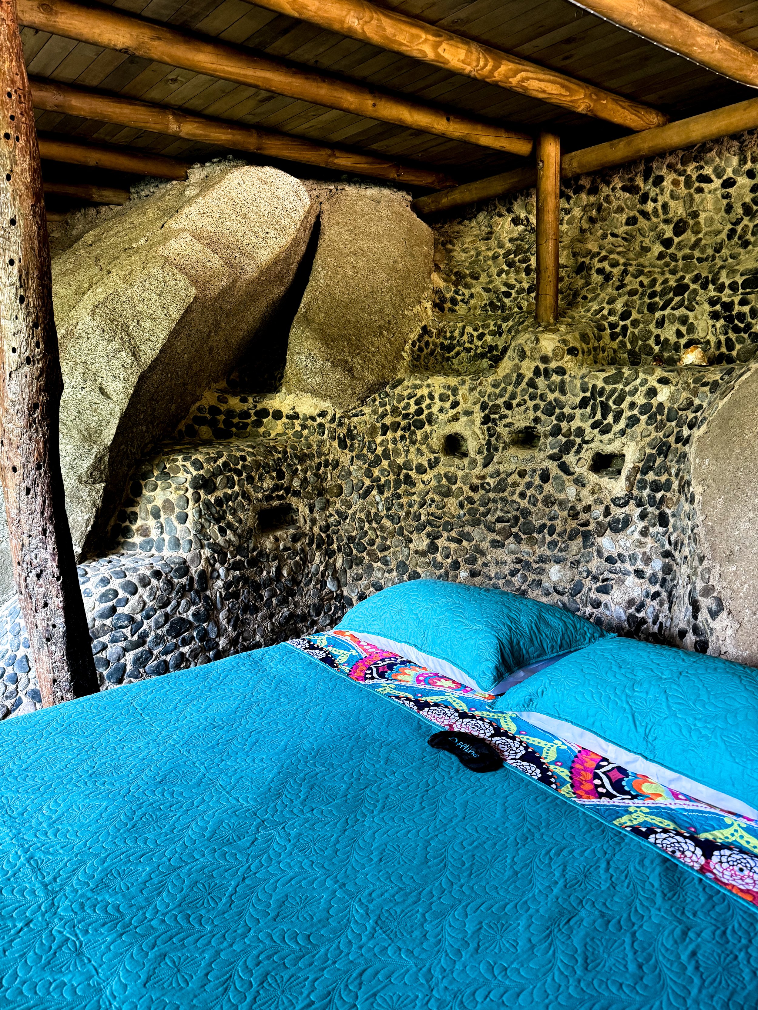 A bedroom with a blue quilted bedspread and colorful fabric strip, against a wall with a large boulder and a pebble mosaic design.