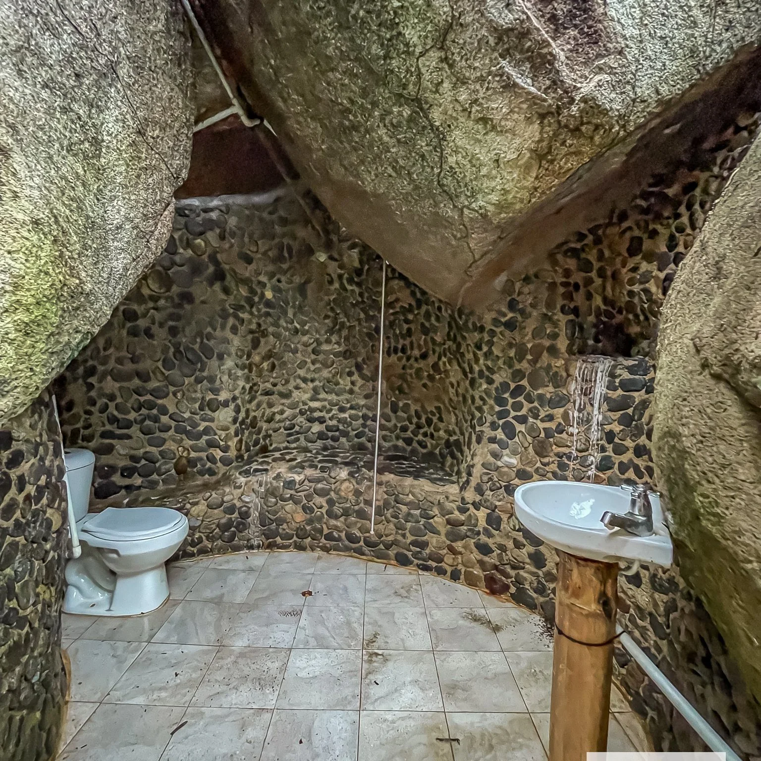 A rustic bathroom with stone walls and ceiling, featuring a toilet and a small sink mounted on a wooden stand, with a tiled floor and a small water feature on the wall.