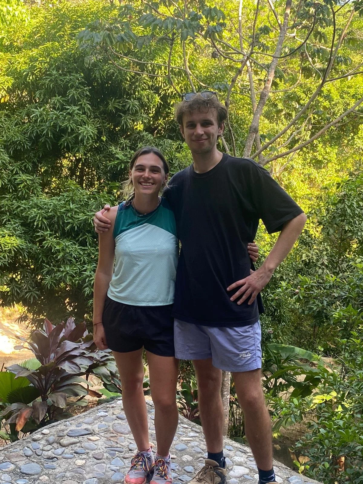 A young woman and man standing close together outdoors in a lush, green garden, smiling at the camera. They are dressed in casual athletic clothing, with the woman in a sleeveless top and shorts and the man in a T-shirt and shorts.