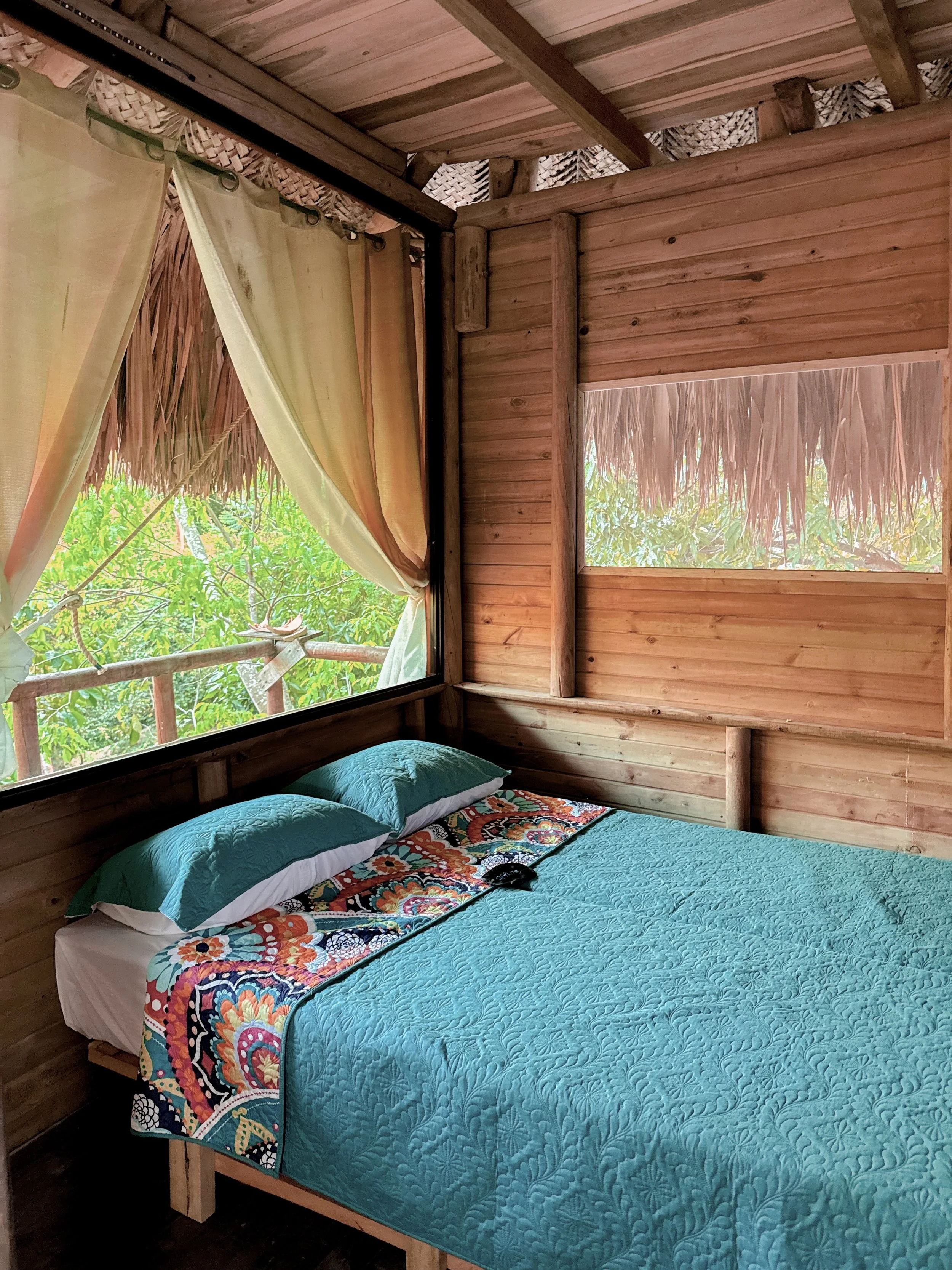 A cozy bedroom with wooden walls and ceiling, featuring a large window with curtains overlooking lush green trees, and a bed with a turquoise quilt, matching pillows, and a colorful blanket.