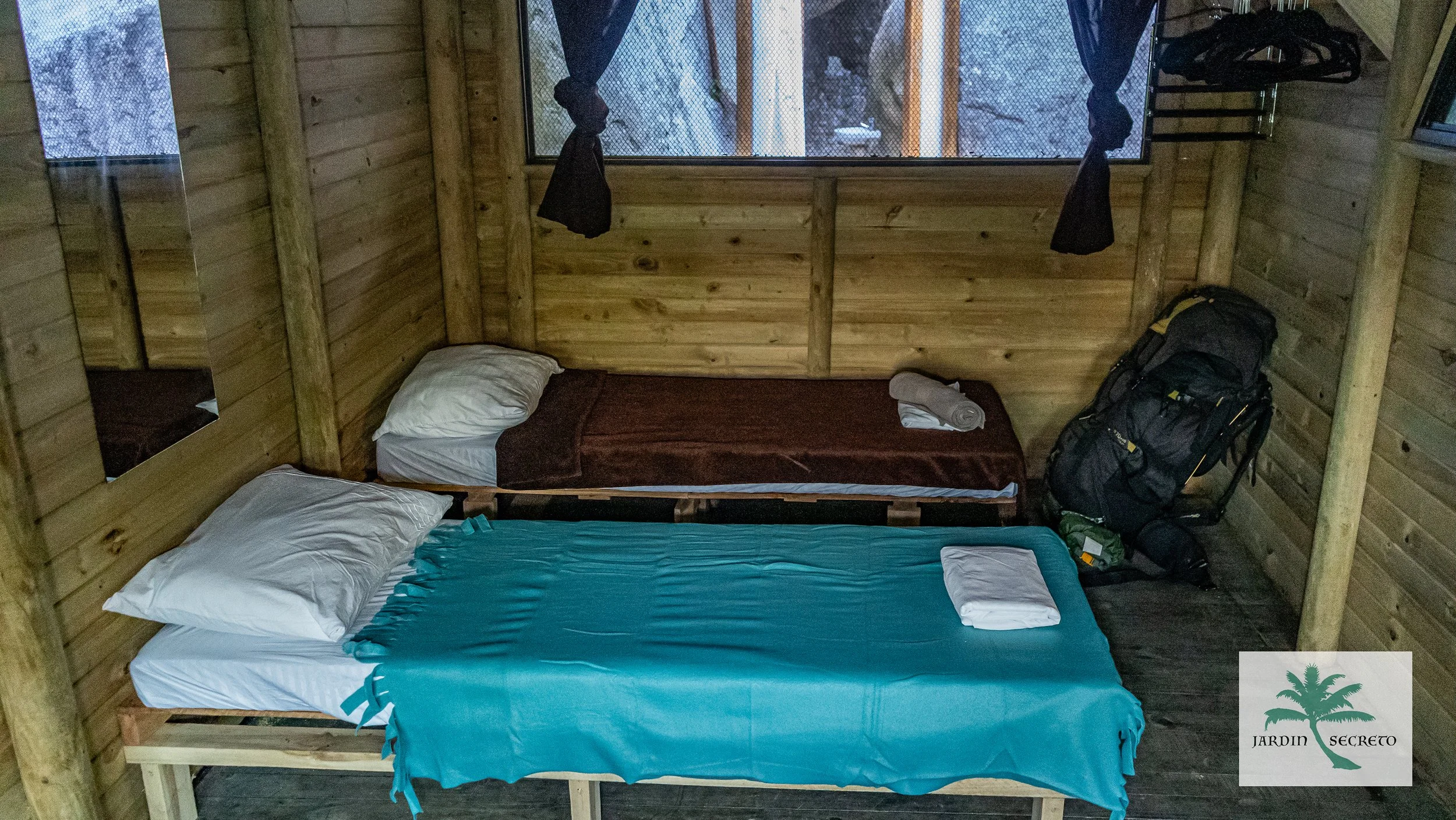 A small wooden cabin room with two beds, one with a blue bedspread and one with a brown blanket, pillows, and towels. There is a window with black curtains and a backpack near the corner.