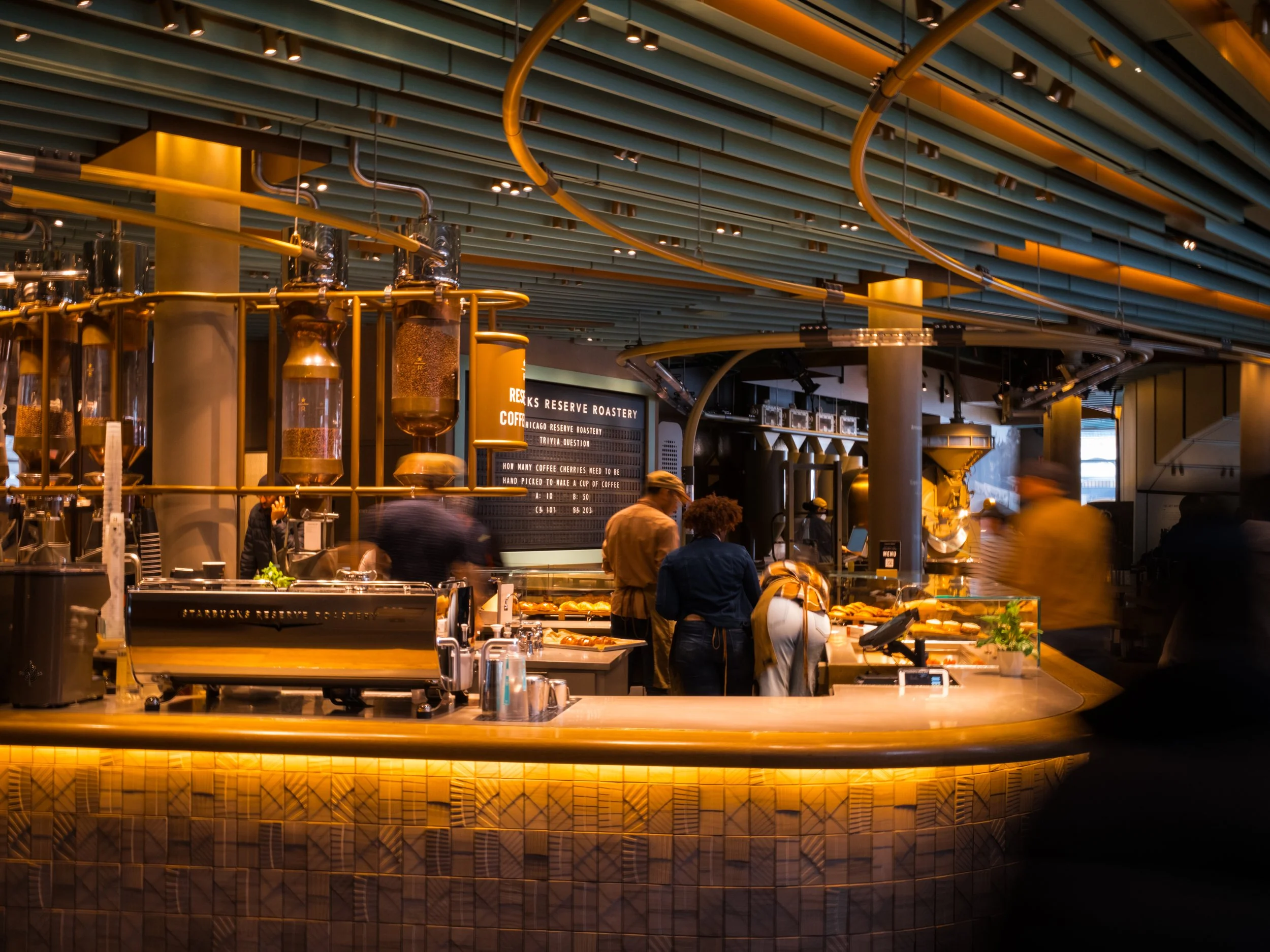 Inside of a modern coffee shop or café with customers ordering and working behind the counter, decorated with warm lighting and industrial design elements.