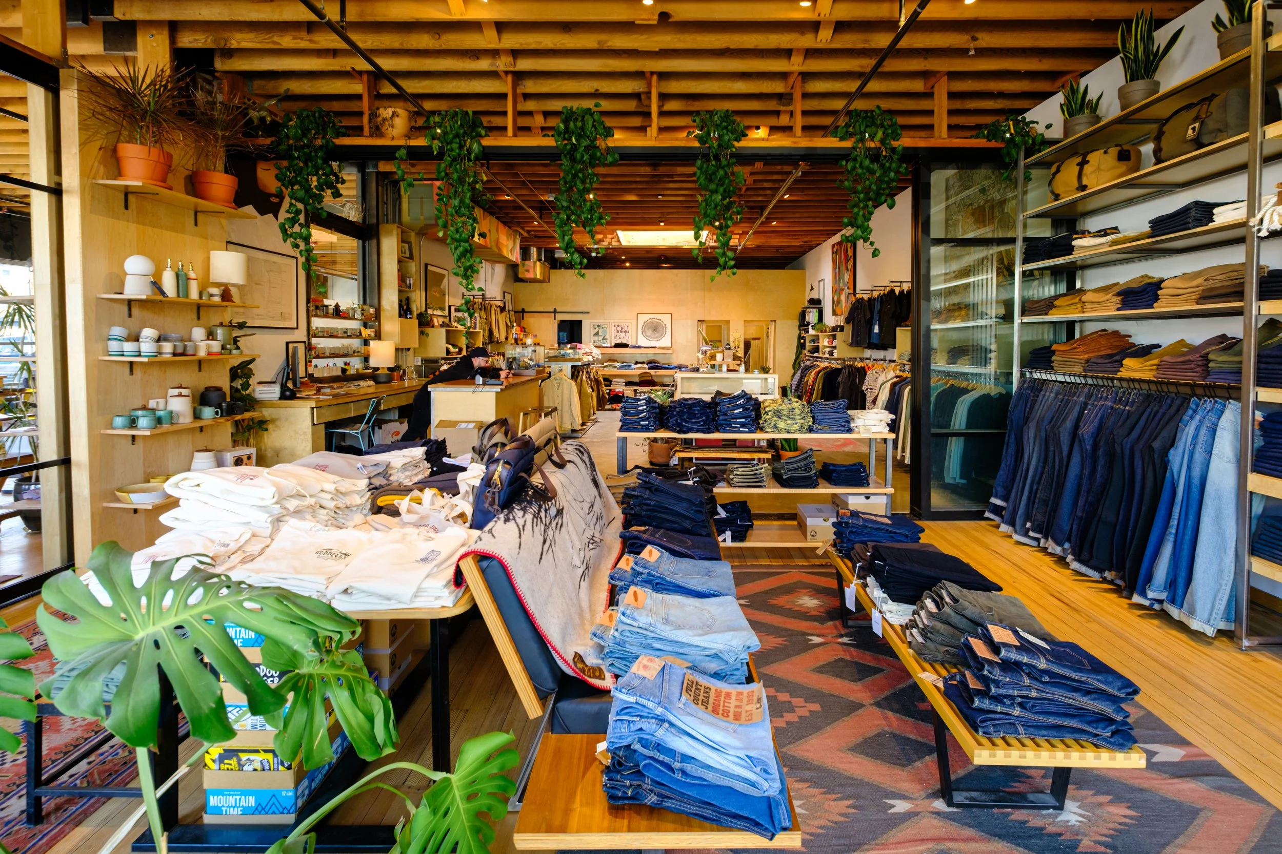 Inside a clothing boutique with racks of jeans and shirts, wooden shelves with plants and accessories, and a patterned rug on the wooden floor.