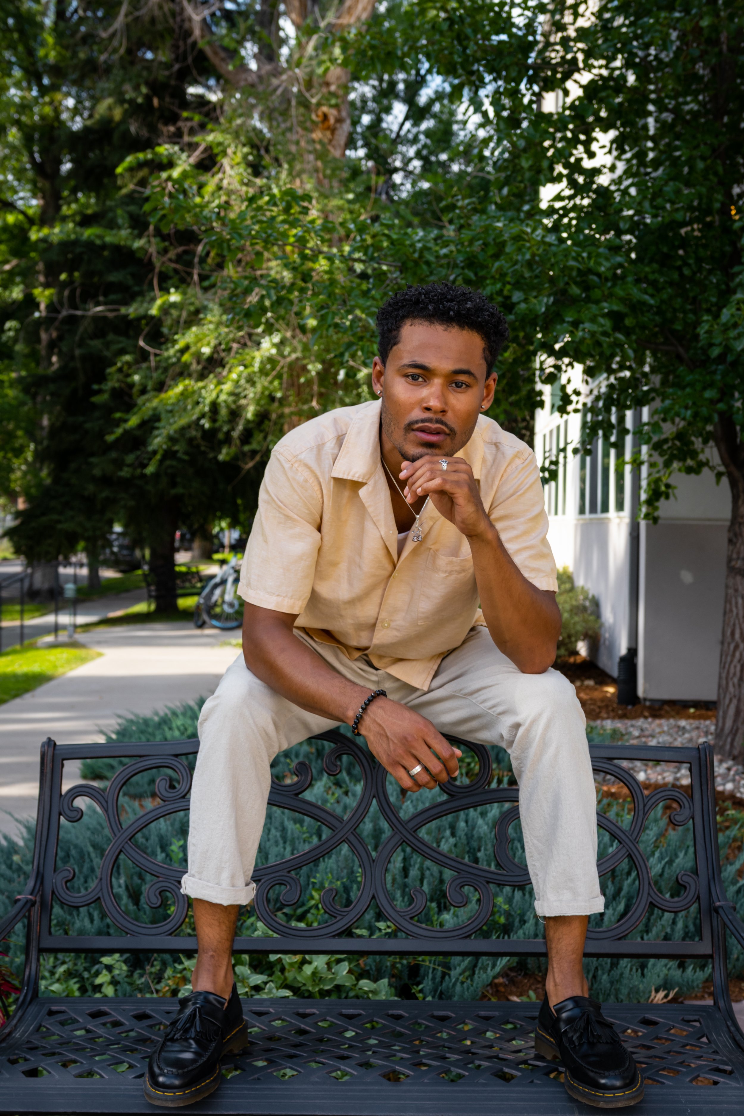 A man with curly black hair and medium brown skin tone sitting on a black ornate metal bench outdoors. He is wearing a beige short-sleeved shirt, cream-colored pants with cuffs, black loafers, and jewelry including rings, a bracelet, a necklace, and 
