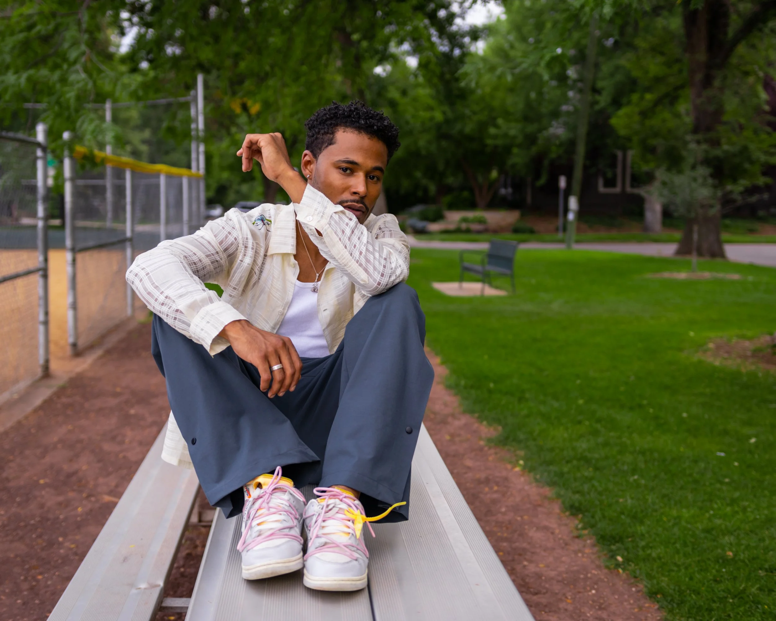 A young man sits on a bench in a park, leaning forward with one arm resting on his knee, wearing a white shirt, dark pants, and colorful sneakers, with greenery and playground fencing in the background.