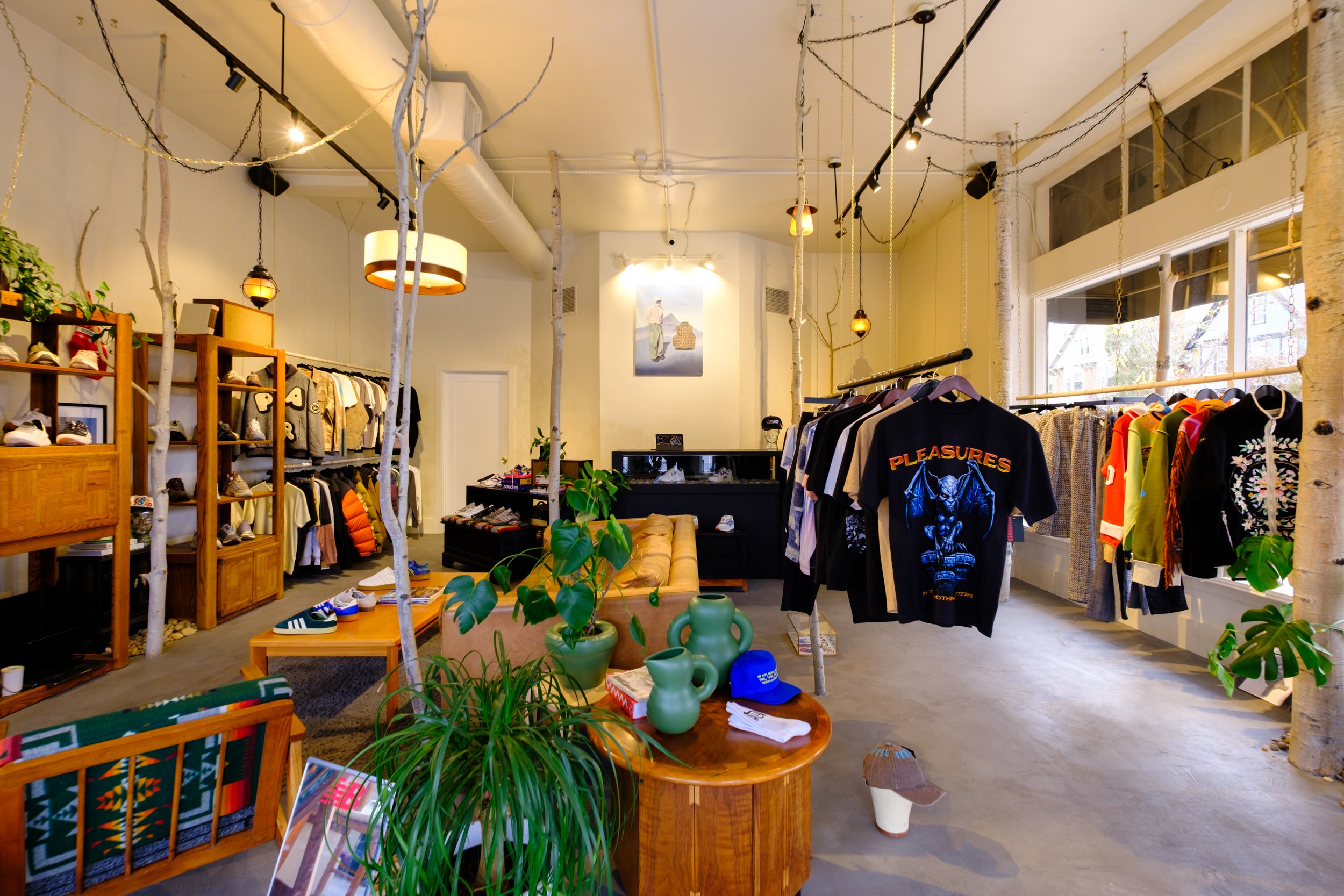 Interior of a boutique clothing store with shelves displaying shoes, racks with t-shirts, jackets, and pants, potted plants, wooden furniture, and decorative lighting.