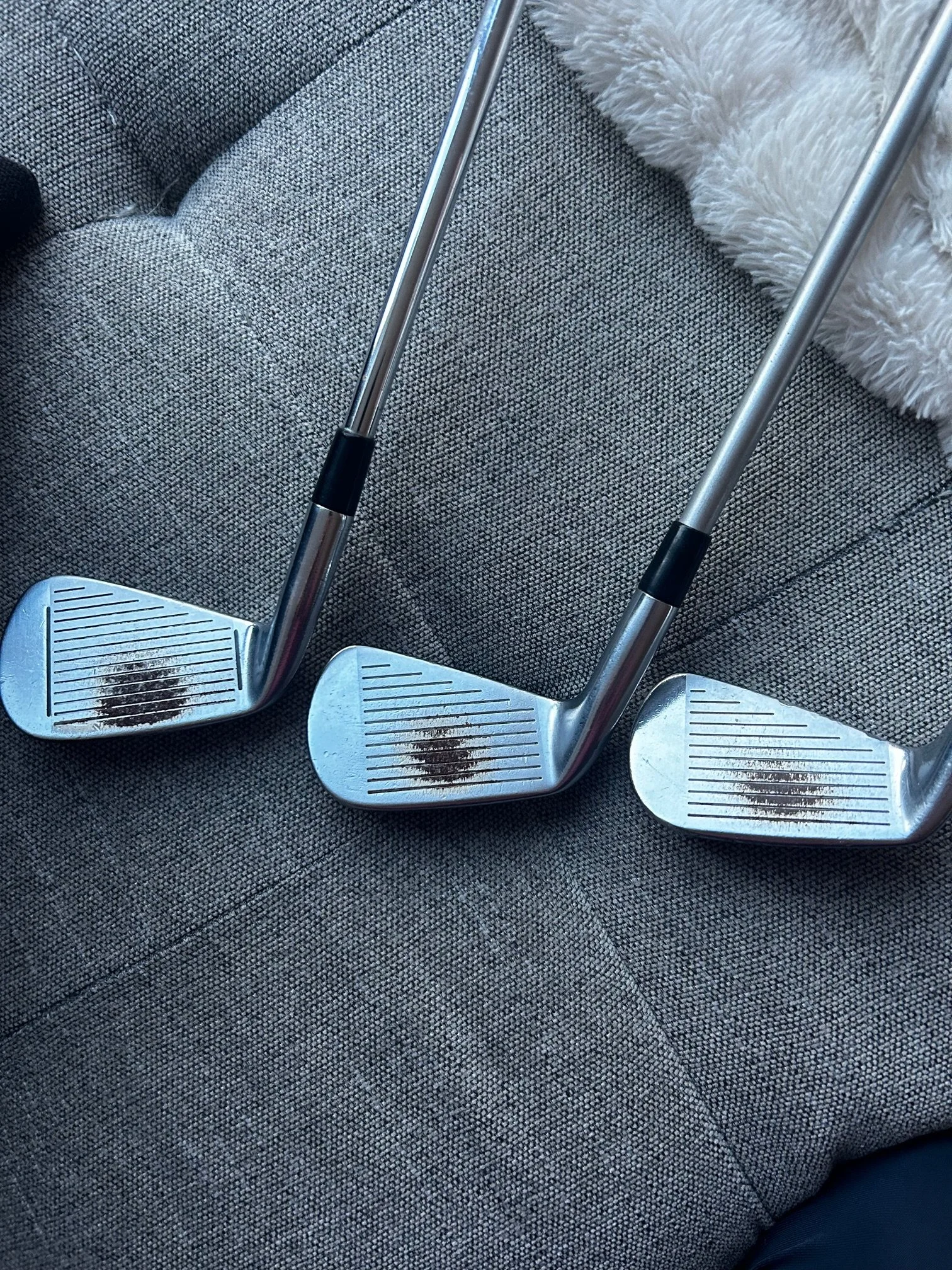 Three golf clubs resting on a gray fabric surface with a fuzzy white blanket nearby.