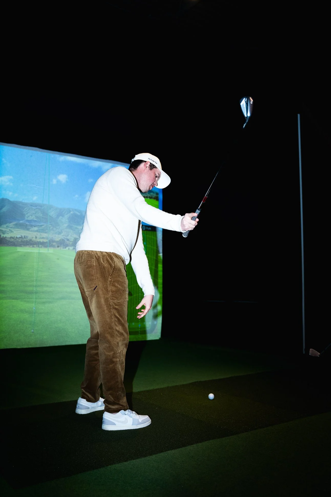 A man in a white sweater, brown pants, and white sneakers is practicing golf indoors on a simulator, swinging a golf club at a golf ball.