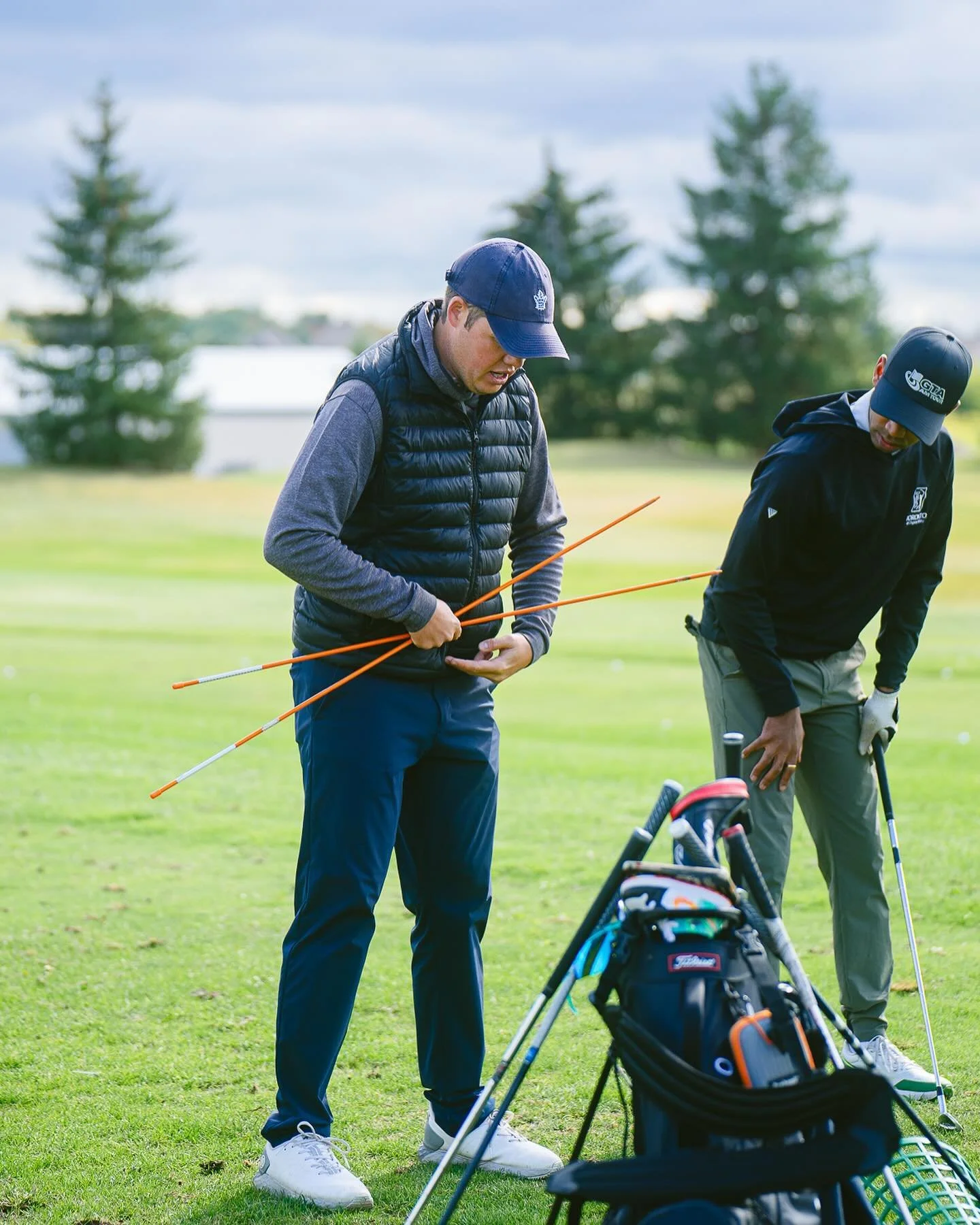 Two men on a golf course preparing to play, one standing and holding two golf club shafts, the other bent over adjusting his golf club, with golf bags and clubs on a cart in front of them, trees and a cloudy sky in the background.