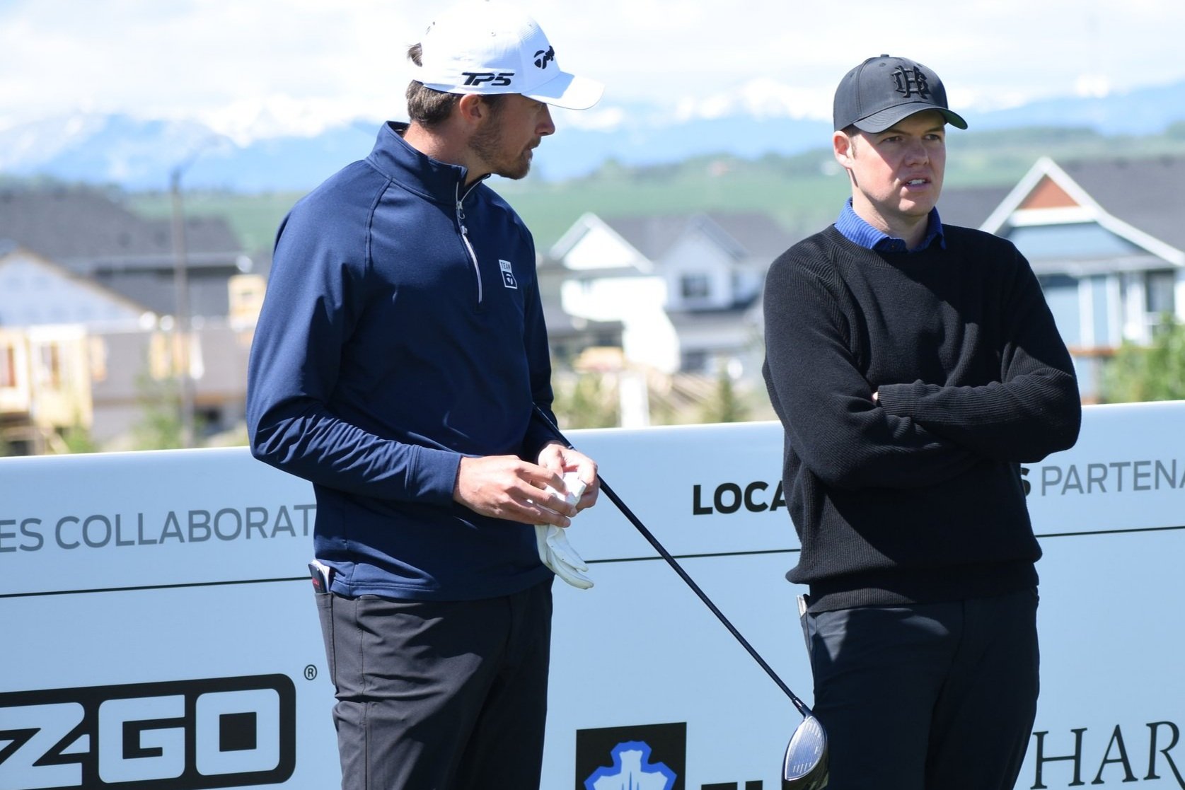 Two men standing outdoors near a golf course, one holding a golf club and the other with arms crossed, with houses and mountains in the background.