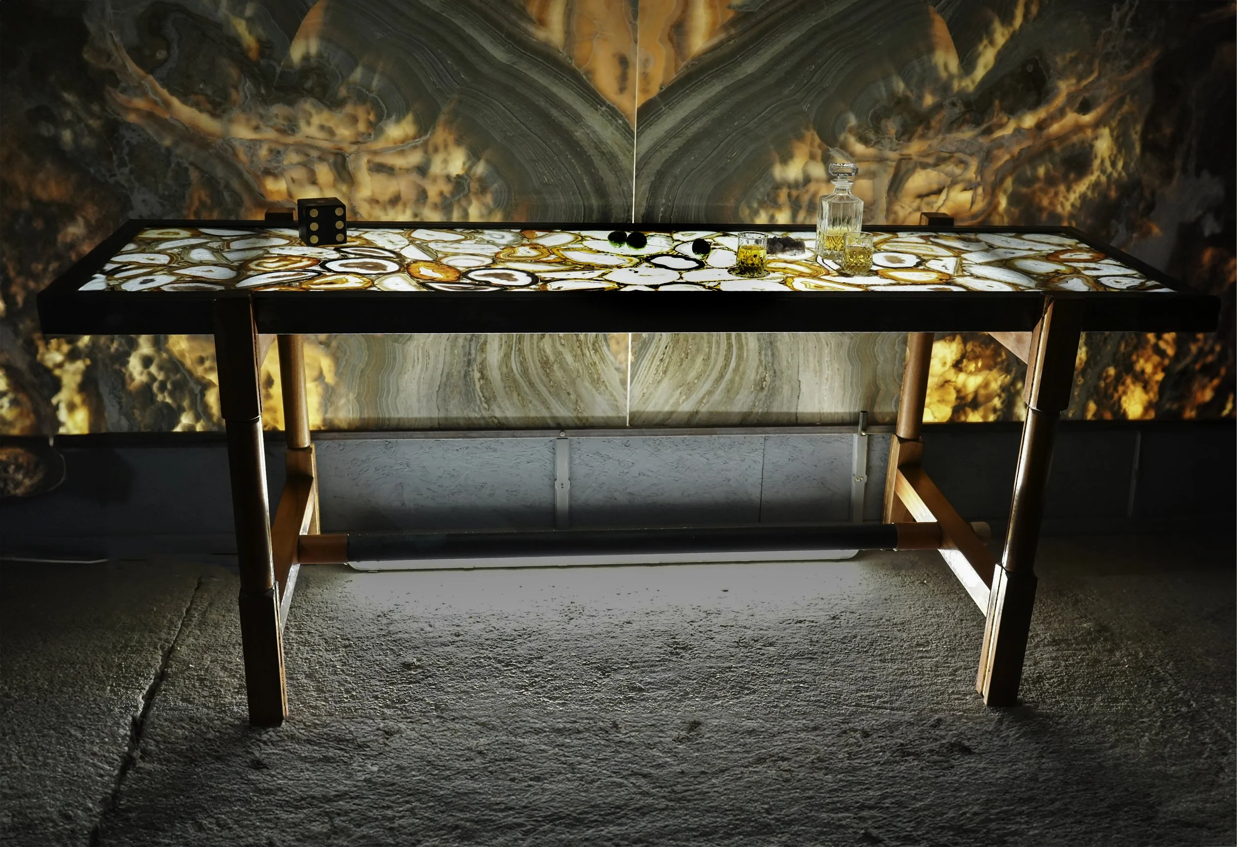 A rectangular table with a top featuring orange, white, and gray swirls, set against a stone or concrete floor. On the table are two small glasses of a yellow liquid, a clear decanter, and some small black and green objects. The background has a large, abstract, multicolored stone or mineral pattern.