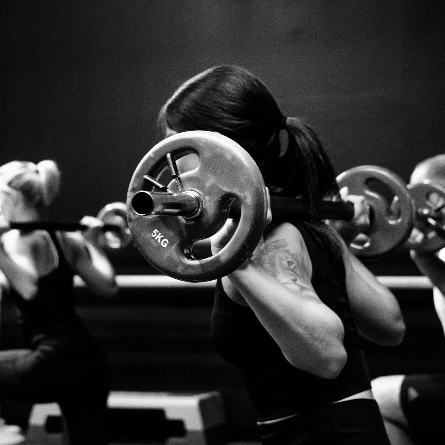 A woman with long dark hair training with a barbell loaded with weight plates in a gym.