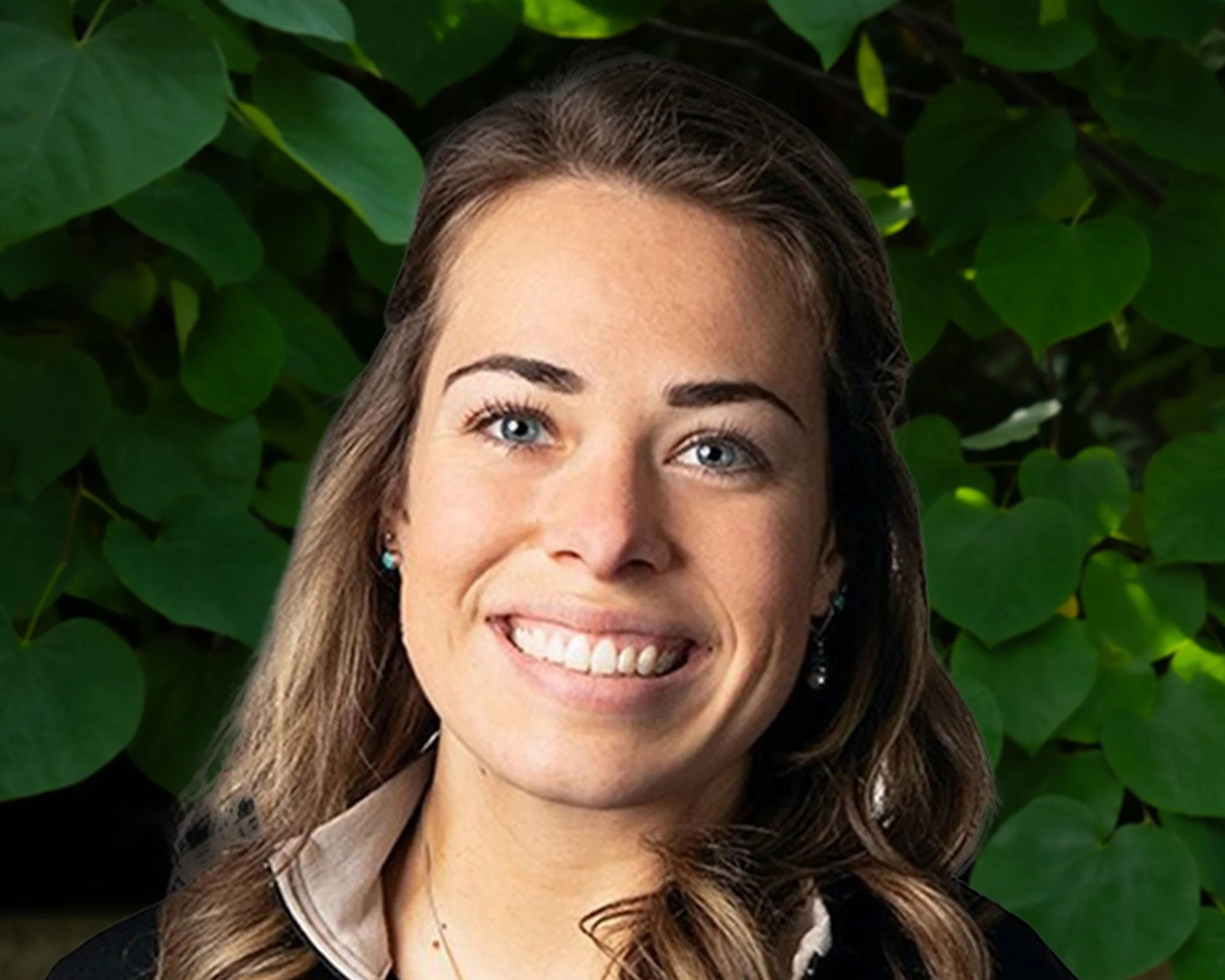 Close-up of a woman with long brown hair, blue eyes, smiling, wearing earrings and a collared shirt, with green leafy background.