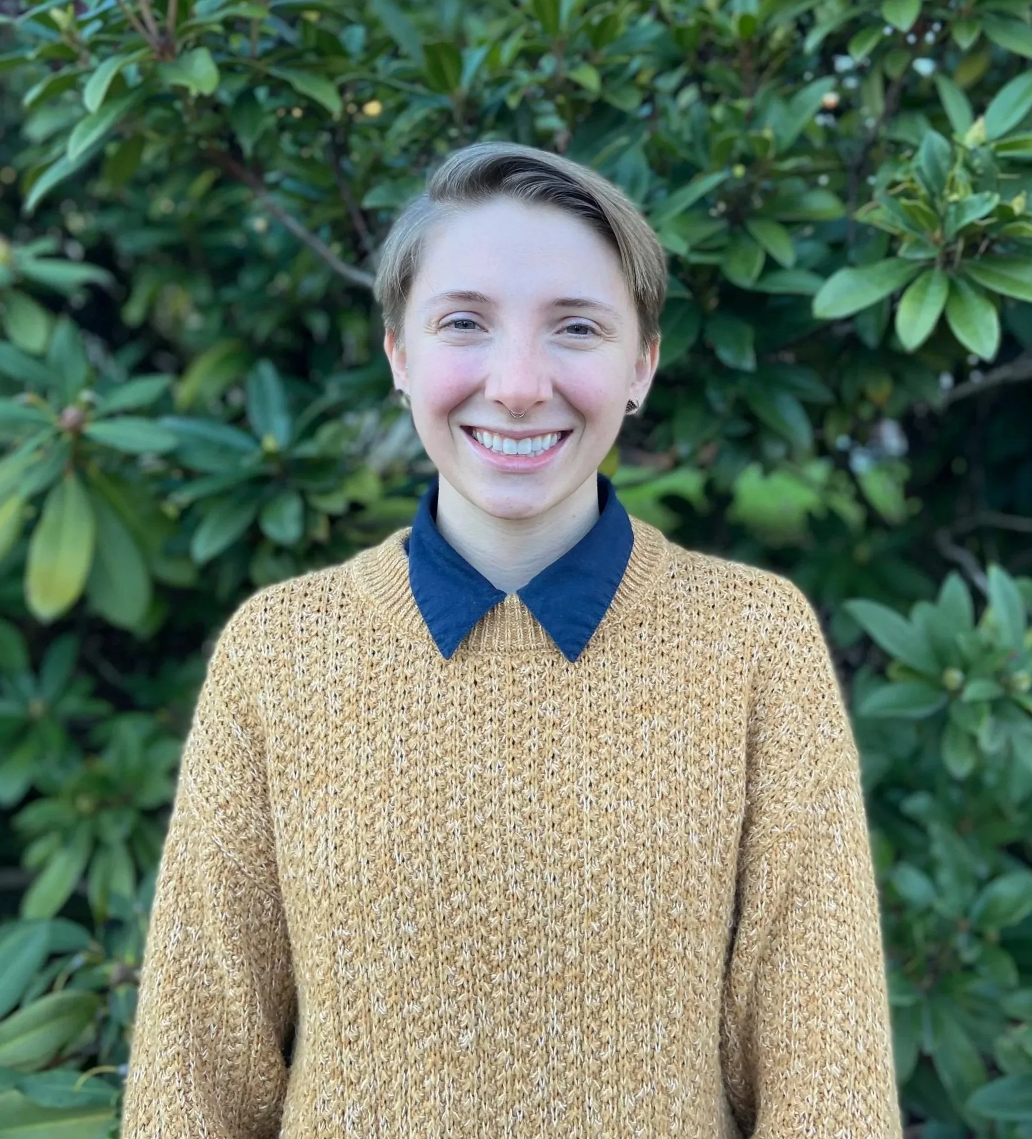 Close-up of a smiling woman with short hair and a septum piercing, wearing a blue shirt and a yellow sweater, standing outdoors in front of green foliage.