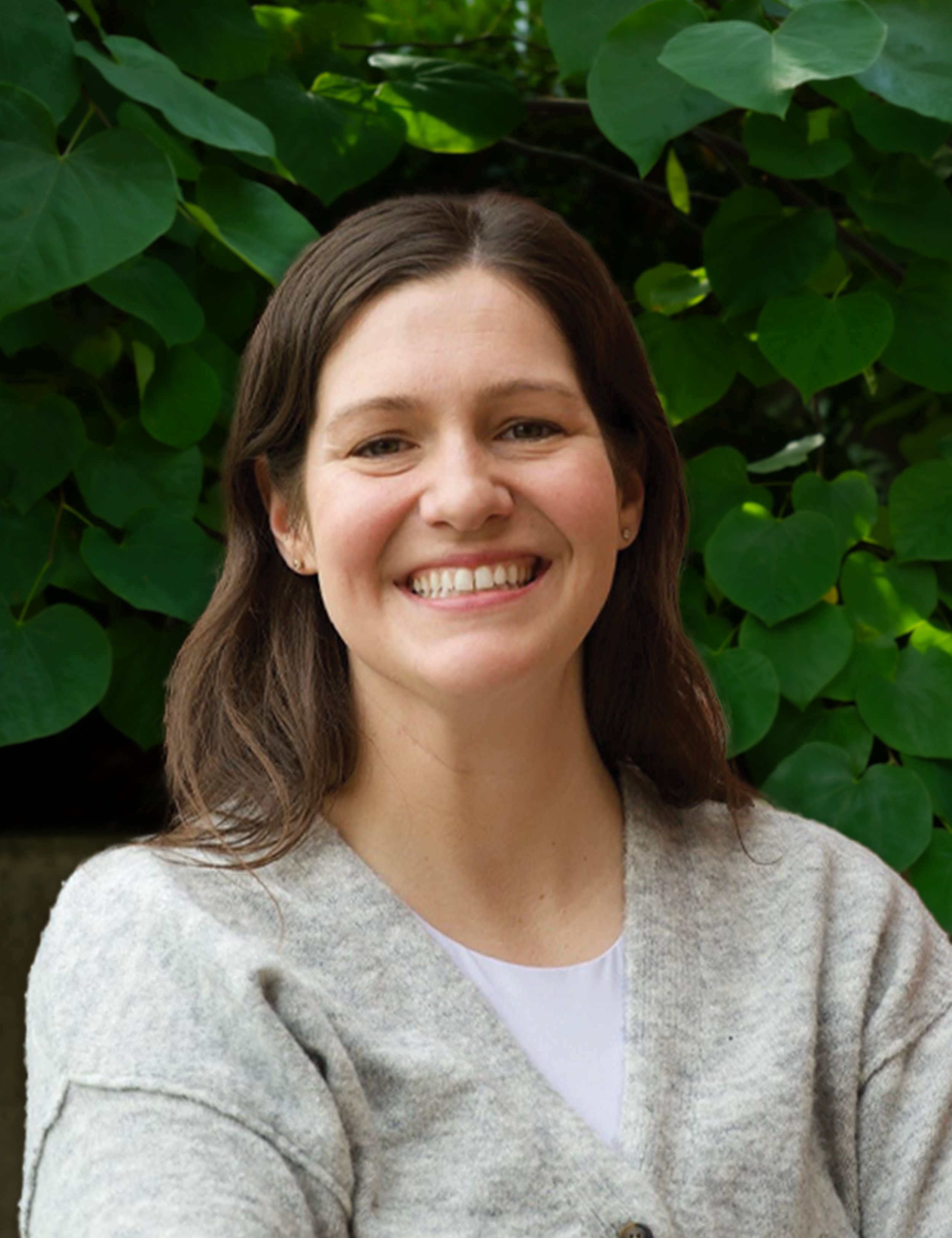 A woman with brown hair, smiling, wearing a light gray cardigan over a white top, in front of green foliage.