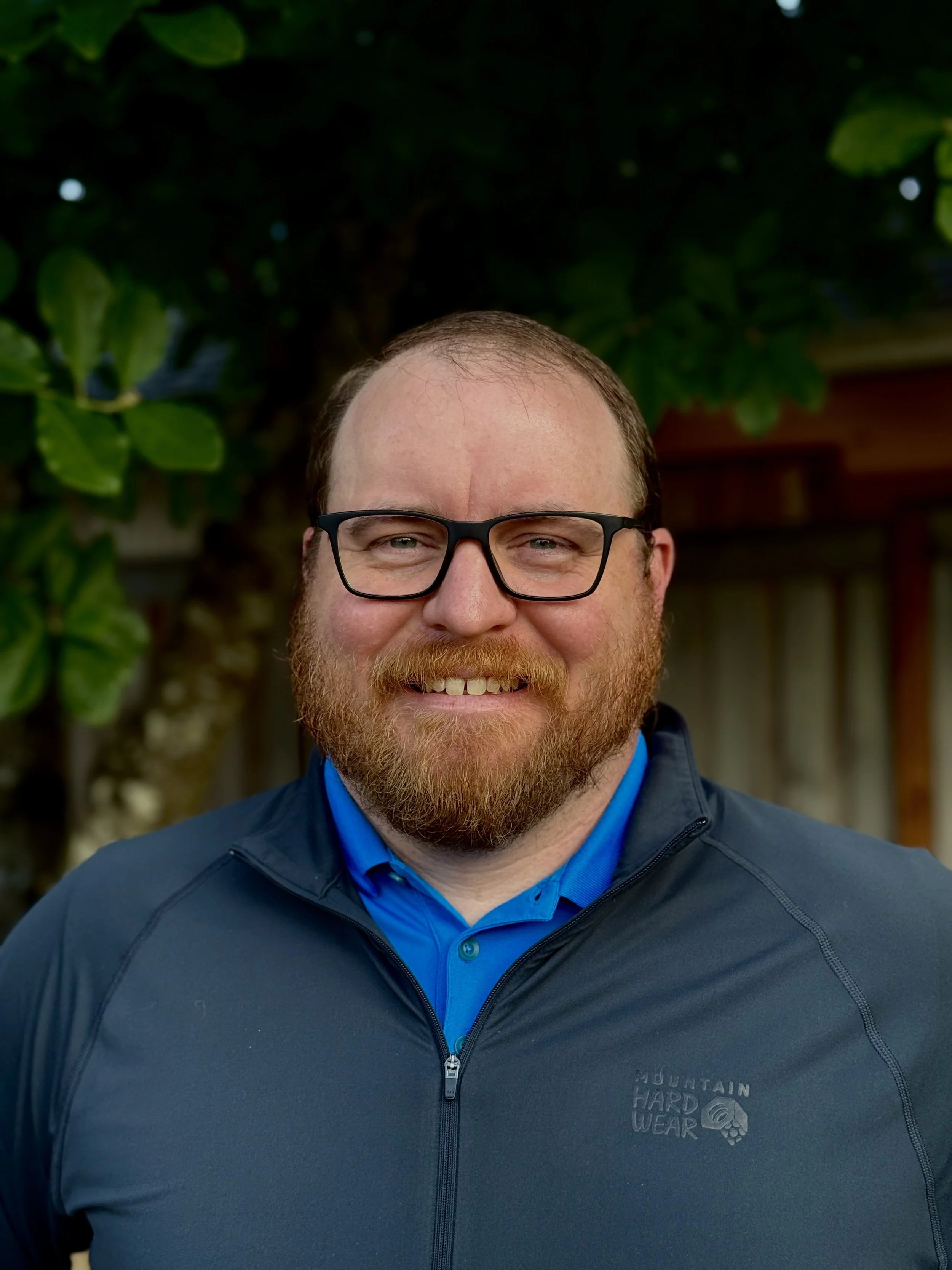 A man with glasses and a beard smiling outdoors in front of trees, wearing a blue collared shirt and a black Mountain Hardwear jacket.
